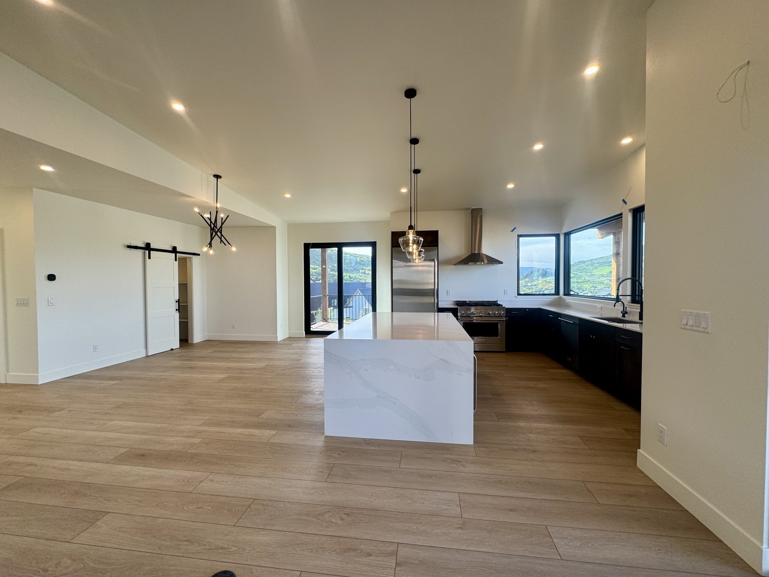 Open-concept kitchen and living area with hardwood floors, white walls, black kitchen cabinets, stainless steel appliances, large windows showing mountain views, and modern lighting fixtures.