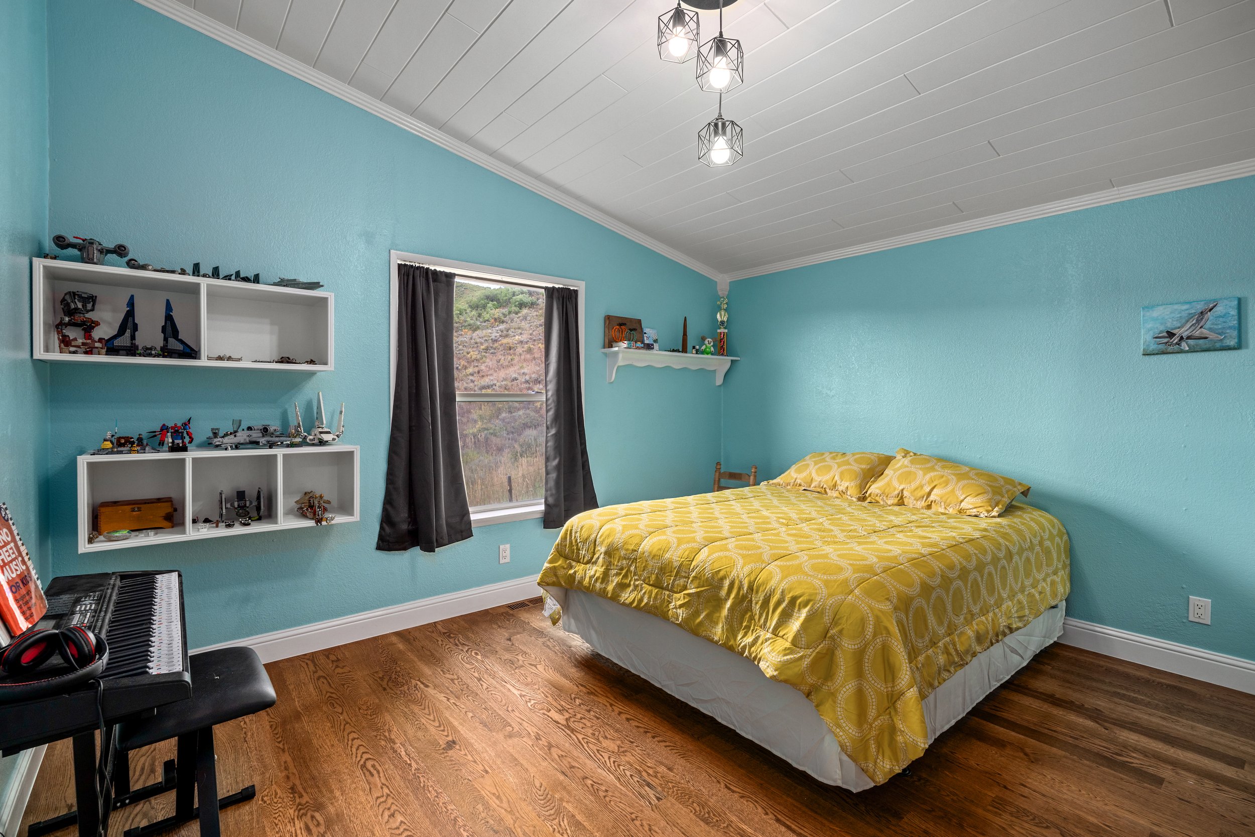 Bedroom with blue walls, yellow-patterned bedding, wooden floor, white shelves with toys and models, a keyboard with headphones, and a window with black curtains.