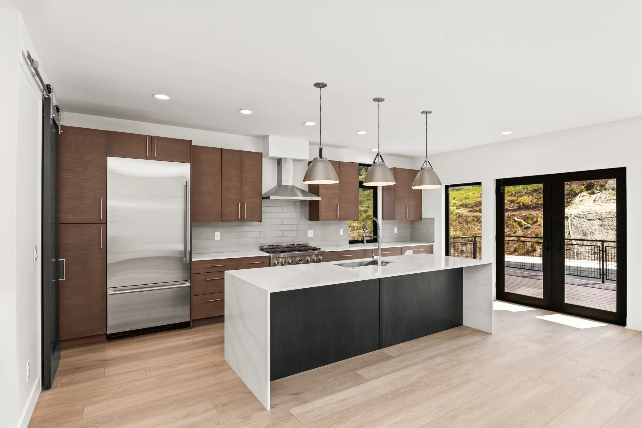 Modern kitchen with brown cabinets, stainless steel appliances, large island, pendant lights, and glass doors.