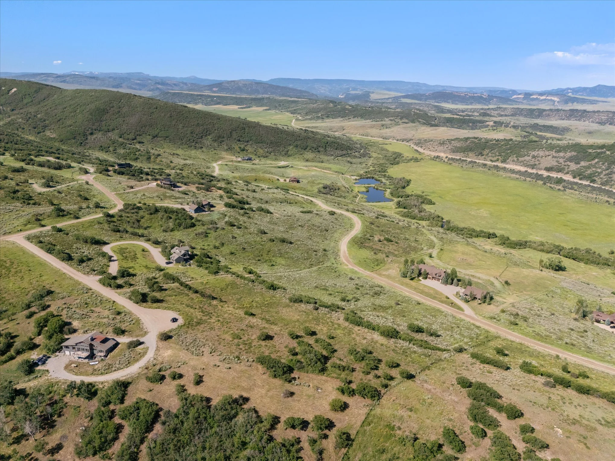 Aerial view of a rural area with winding dirt roads, houses, green fields, a small pond, over rolling hills and mountains in the background.