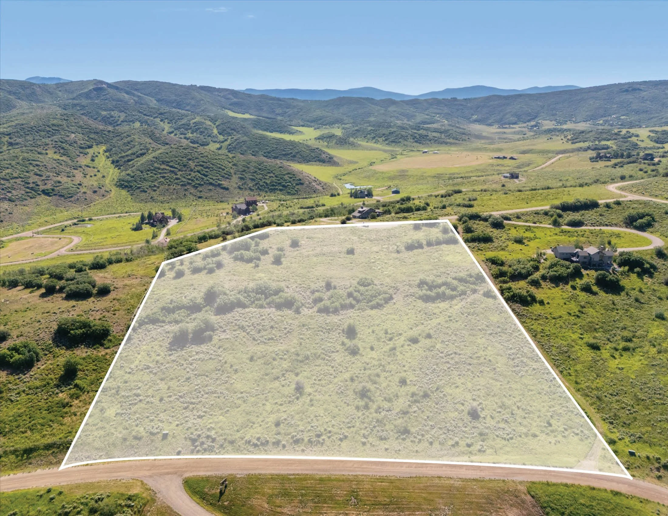 Aerial view of a rural landscape with rolling green hills and a road leading to a large vacant plot of land marked with white boundary lines.