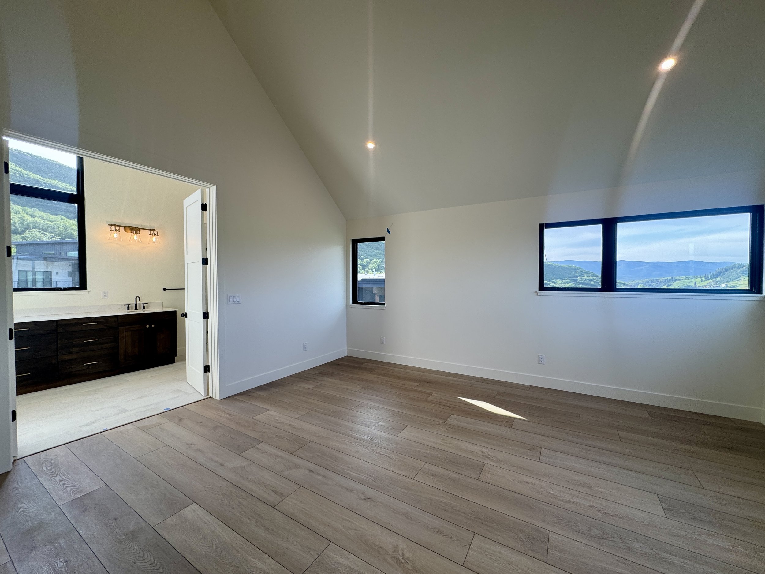Empty room with wood flooring, white walls, large windows showing scenic mountains, and an adjoining bathroom with dark vanity and vanity mirror.