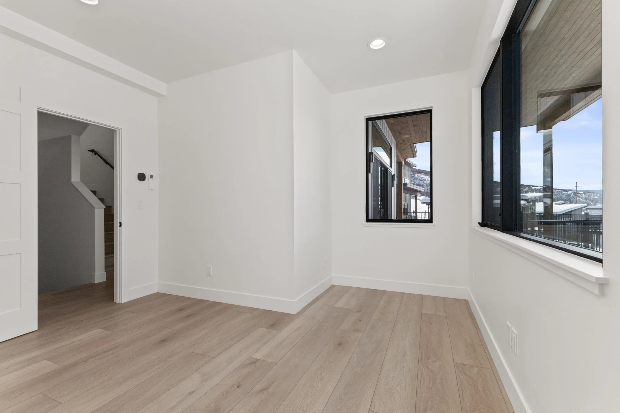 Empty white room with light wood flooring, large windows, and an open doorway leading to a staircase.