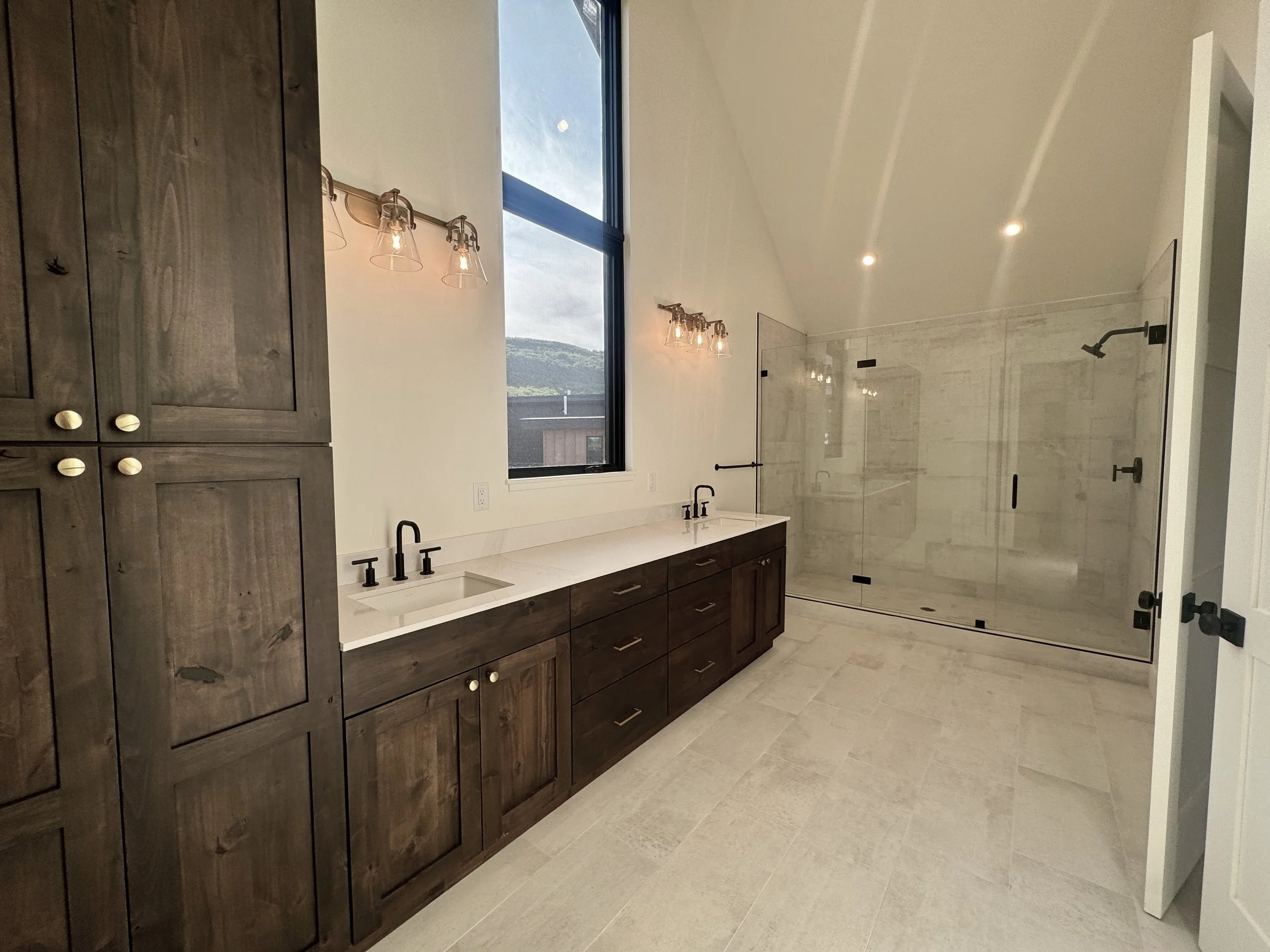Modern bathroom with double vanity, wooden cabinets, marble countertop, black fixtures, large shower with glass enclosure, and light-colored tiled floor.