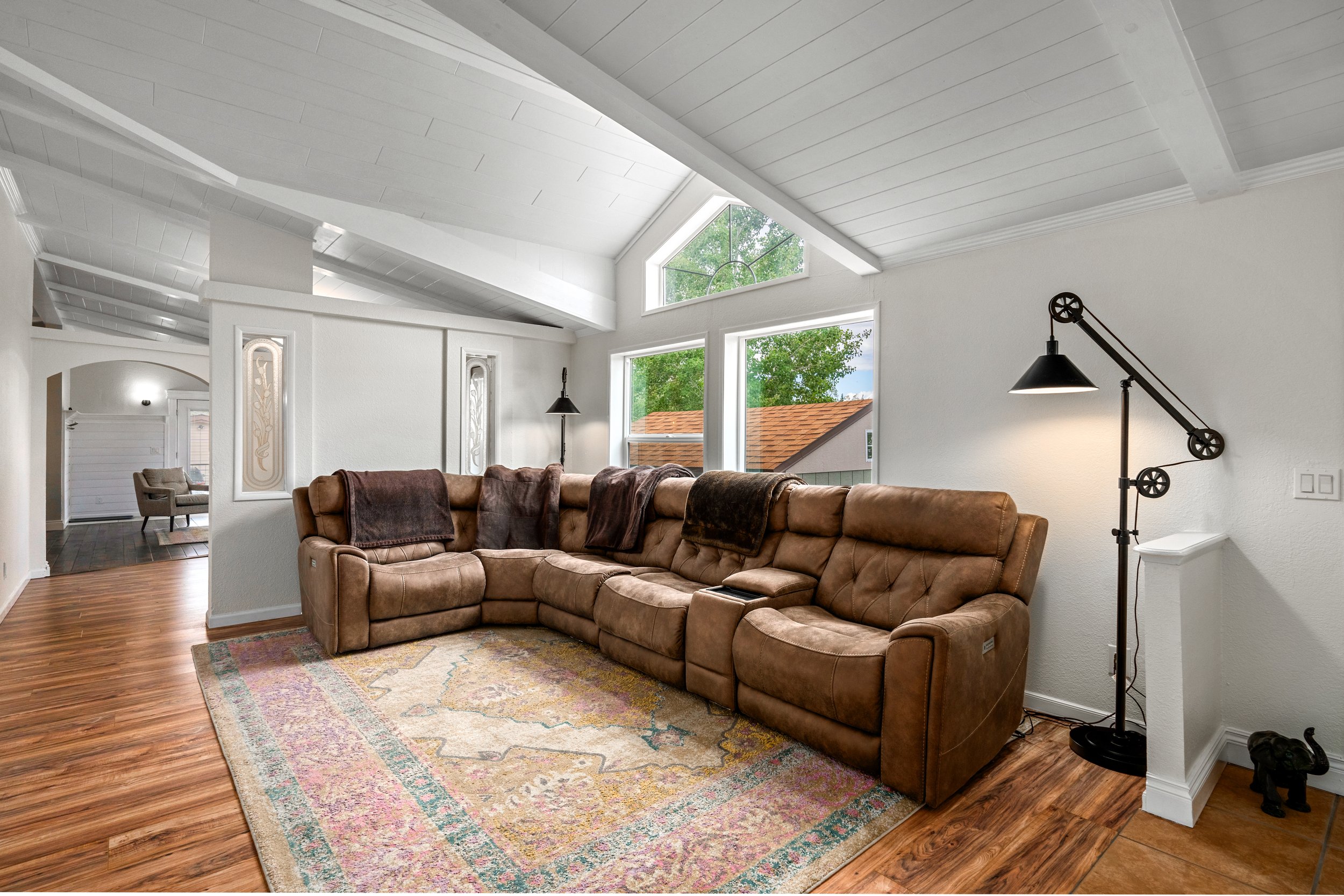 Cozy living room with brown sectional sofa, wooden flooring, area rug, large windows, vaulted white ceiling, and adjustable floor lamp.