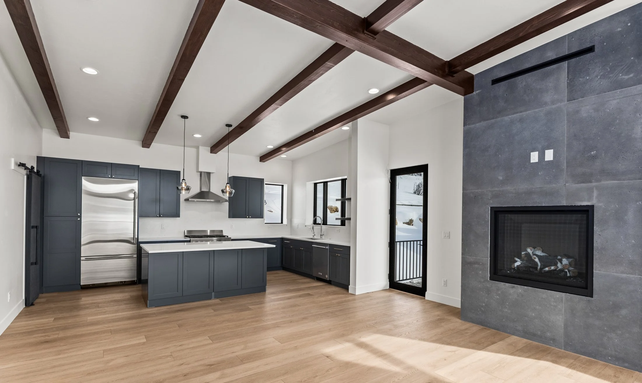 Modern kitchen interior with wooden beams, gray cabinets, stainless steel appliances, and a fireplace.