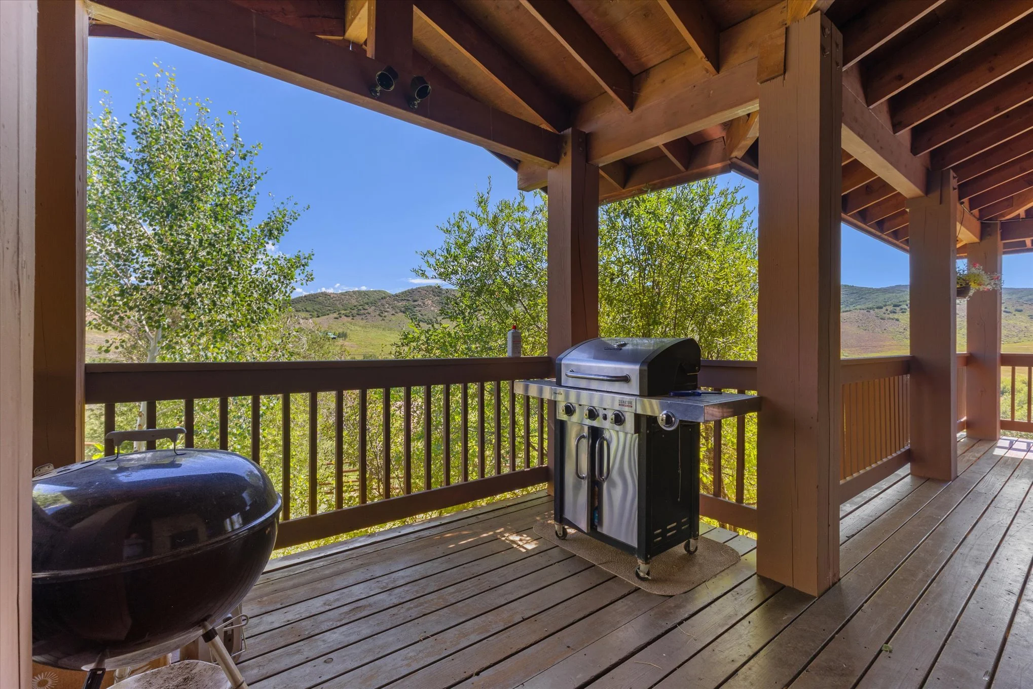 Deck with a barbecue grill and a smoker, overlooking green trees and hills under a blue sky.