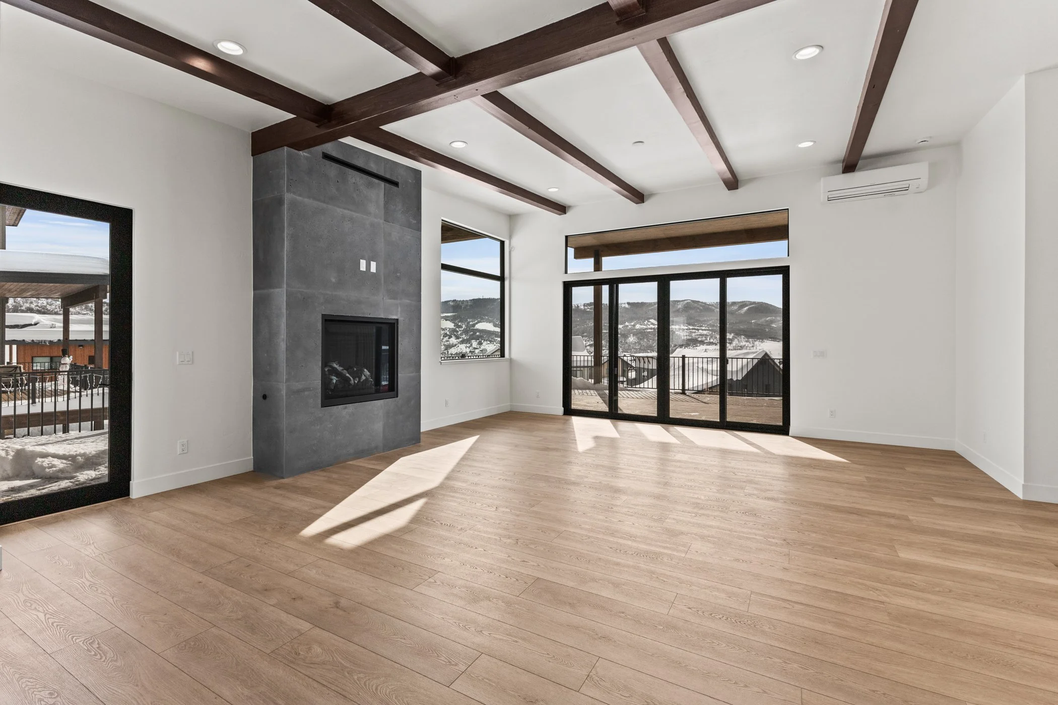 Modern living room with wood beam ceiling, large windows, glass doors, gray fireplace, and light wood floors.