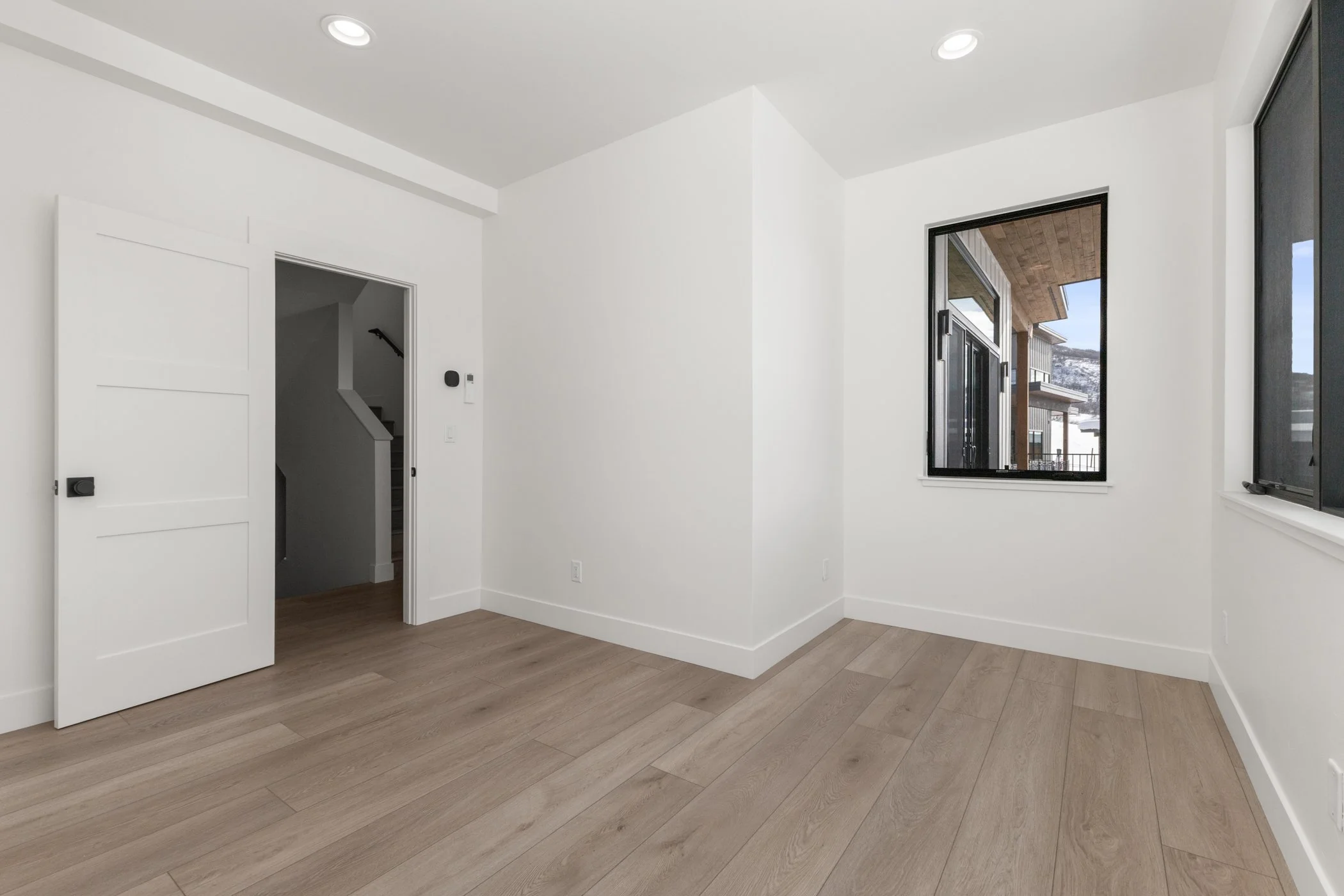 Empty room with light wood flooring, white walls, and a black-framed window showing an outdoor view. There's an open door leading to stairs.