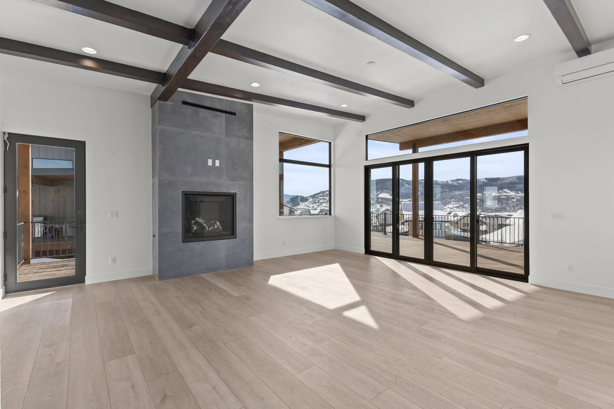 Spacious modern living room with wooden floors, a large window, and a sliding glass door leading to a balcony with a mountain view. The room features a fireplace and dark ceiling beams.