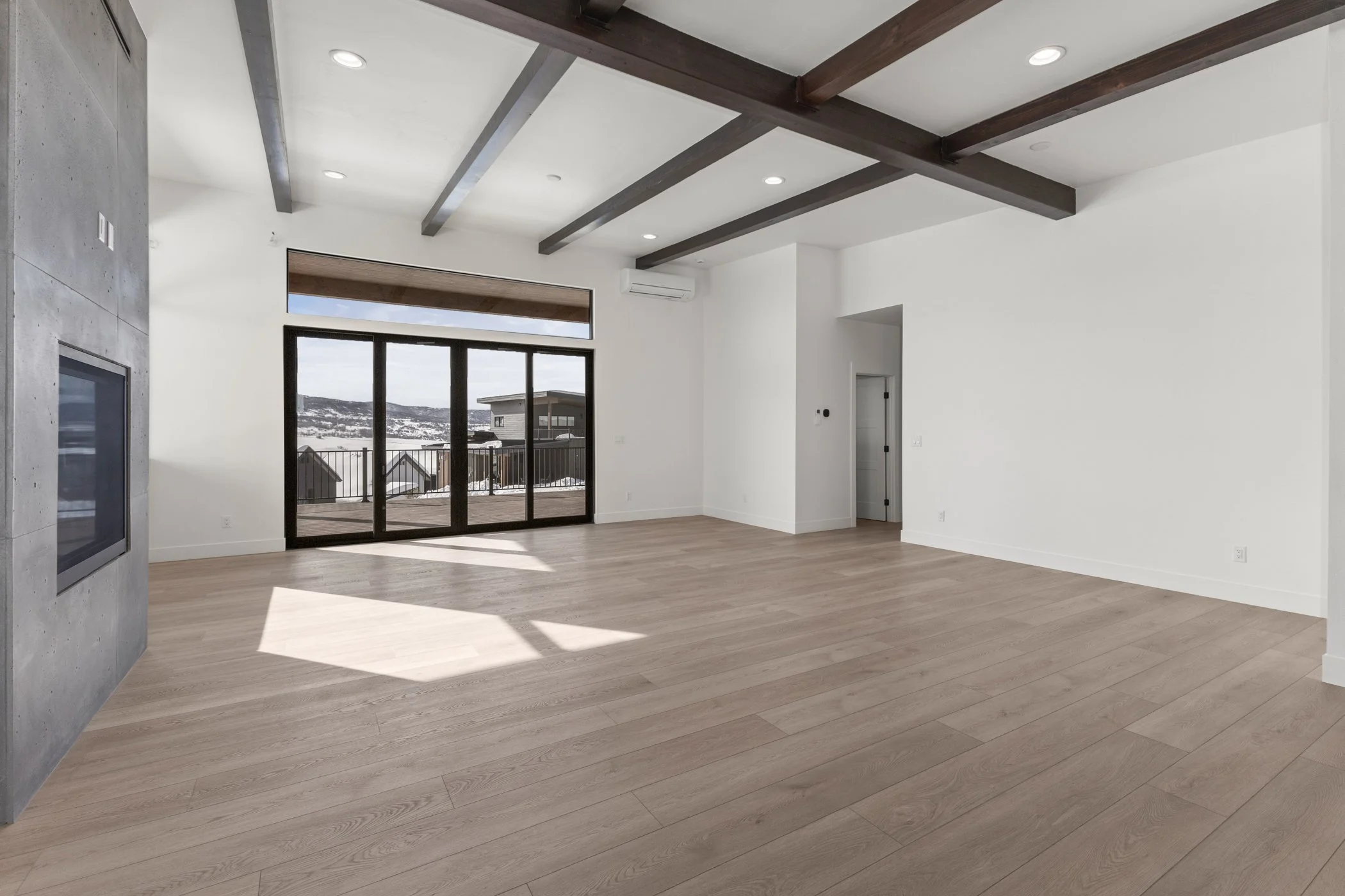 Spacious empty living room with wooden flooring, large windows, exposed beams, and modern fireplace.