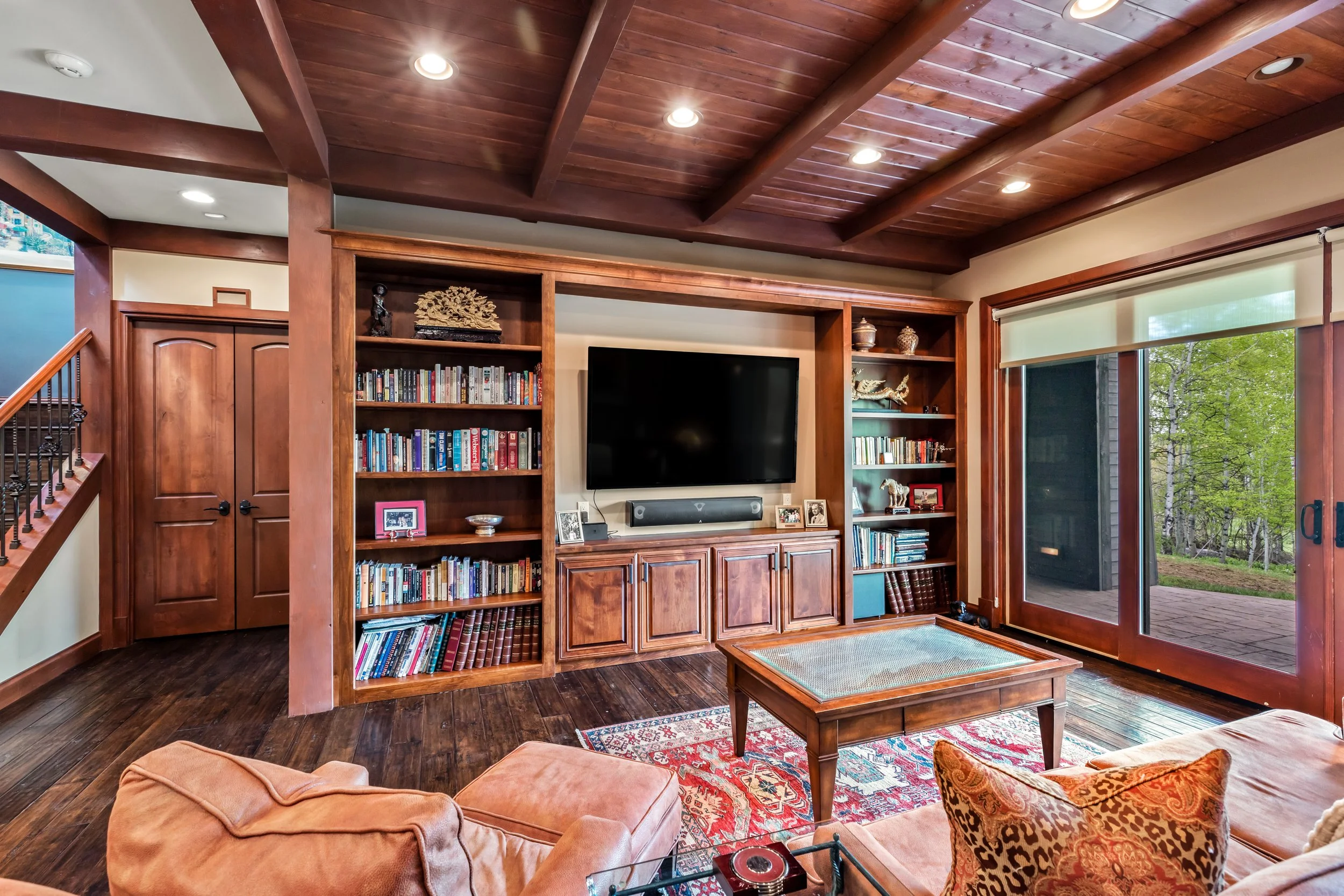 Living room with large wood entertainment center, mounted flat-screen TV, bookshelves, glass sliding door with view of greenery outside, wooden ceiling, hardwood floors, and orange upholstered furniture.