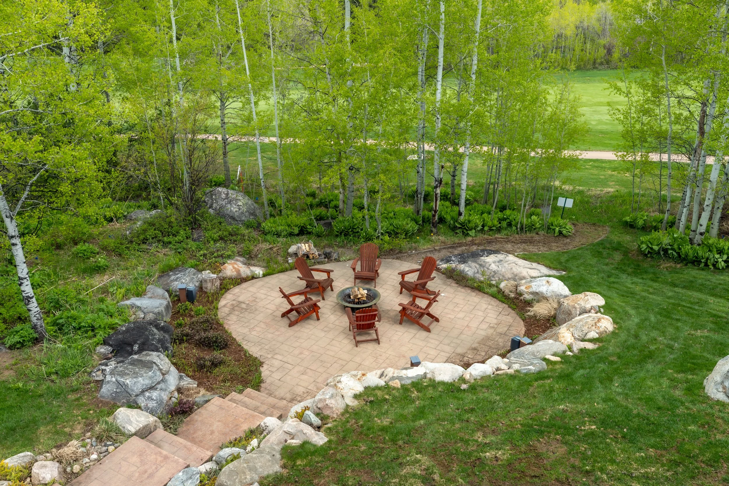 A circular outdoor patio with six wooden Adirondack chairs arranged around a fire pit, surrounded by large rocks and lush greenery, including tall trees and bushes.