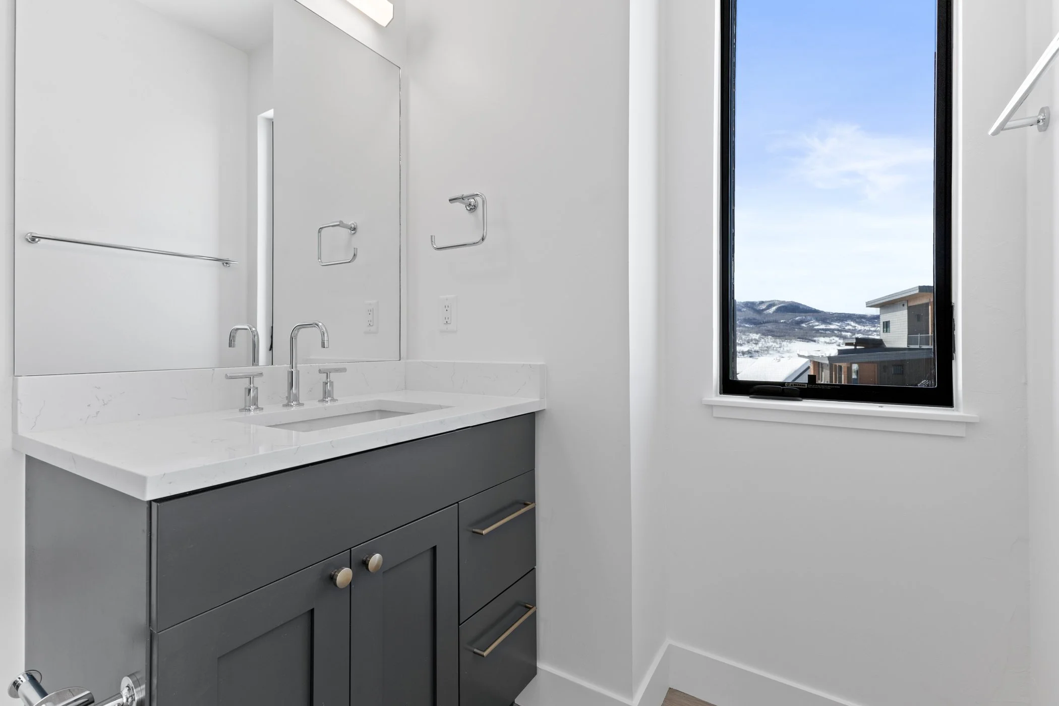 Minimalist bathroom with a gray vanity, marble countertop, large mirror, chrome faucets, and a window showing a snowy mountain view.