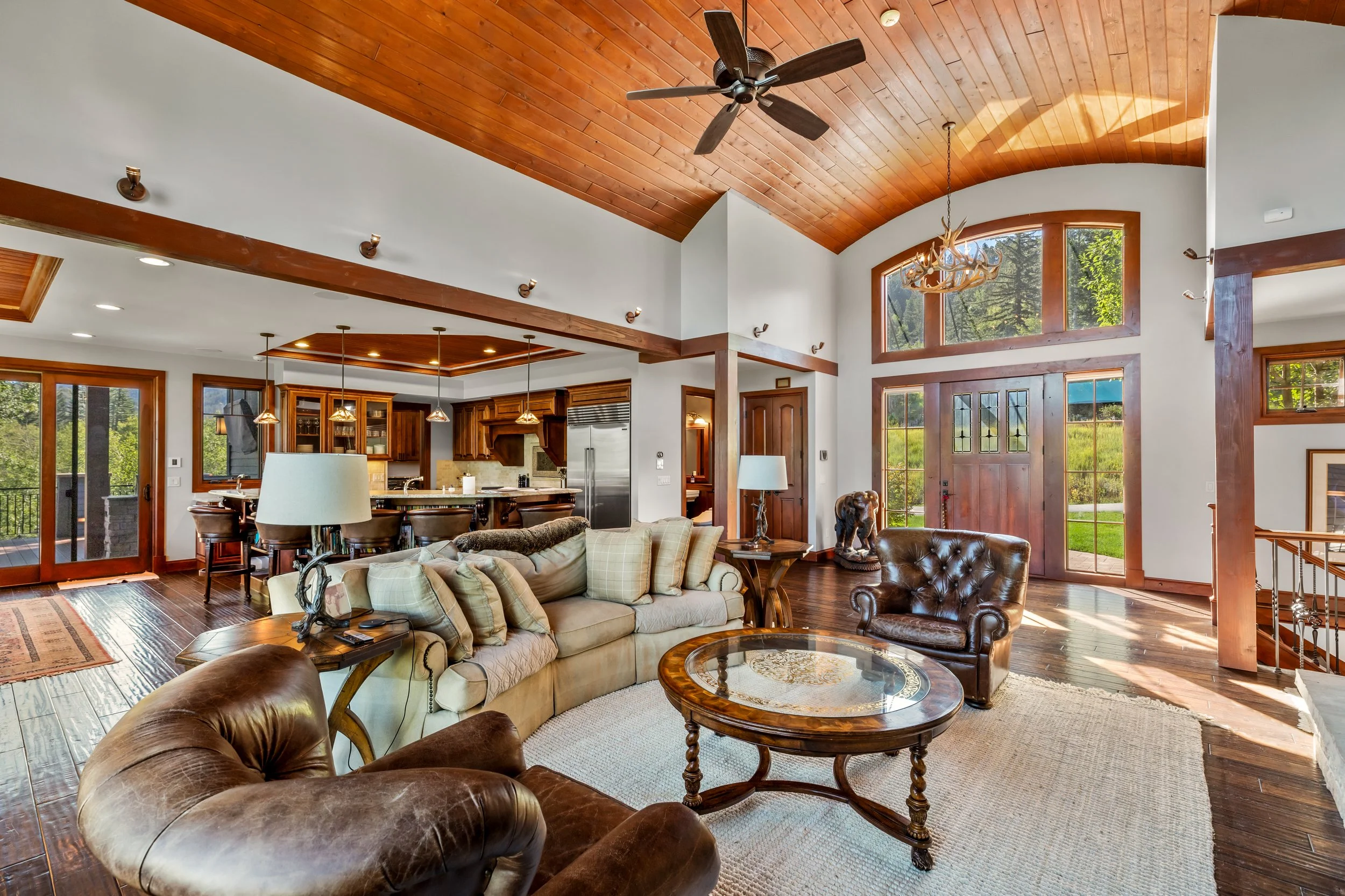Spacious living room with high arched wood-paneled ceiling, large front door with stained glass, multiple windows letting in natural light, and a mix of leather and fabric furniture, including a beige sofa, brown leather armchairs, a wooden coffee ta