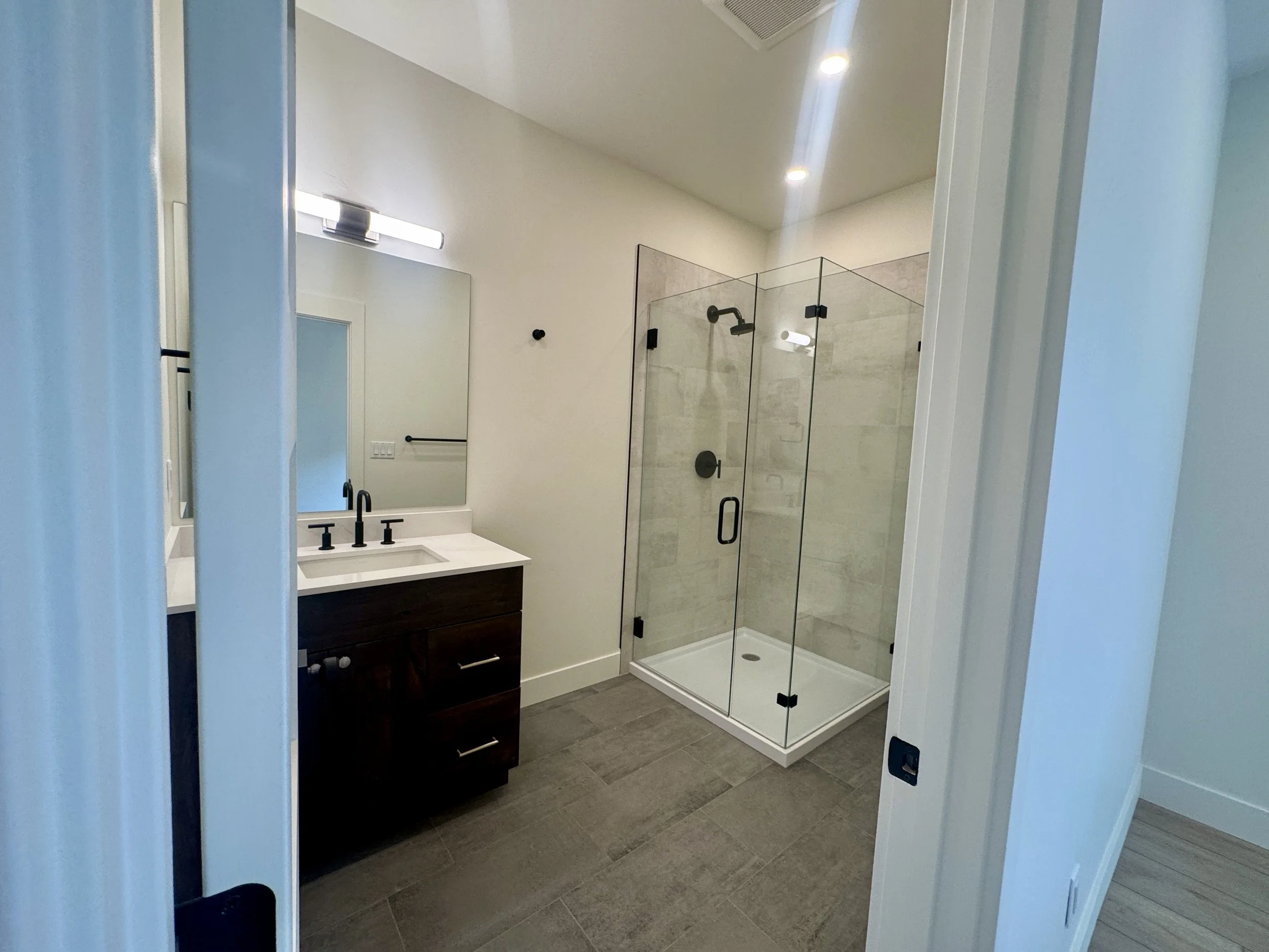 Modern bathroom with a dark wood vanity, a white countertop, a large mirror, a black faucet, and a glass-enclosed shower with beige tiles.