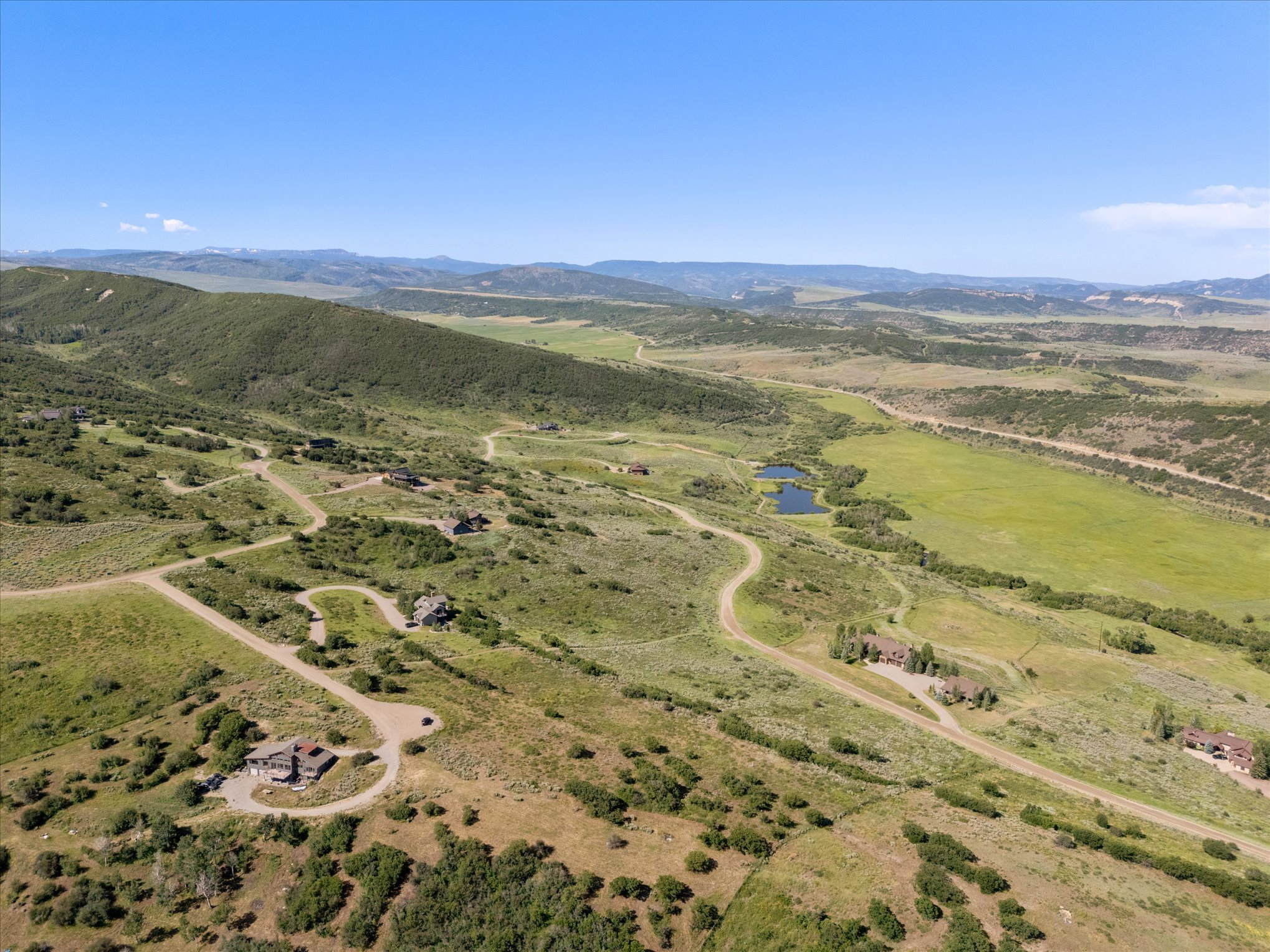 Aerial view of a rural landscape with winding roads, scattered houses, green hills, a small lake, and mountains in the distance under a clear blue sky.