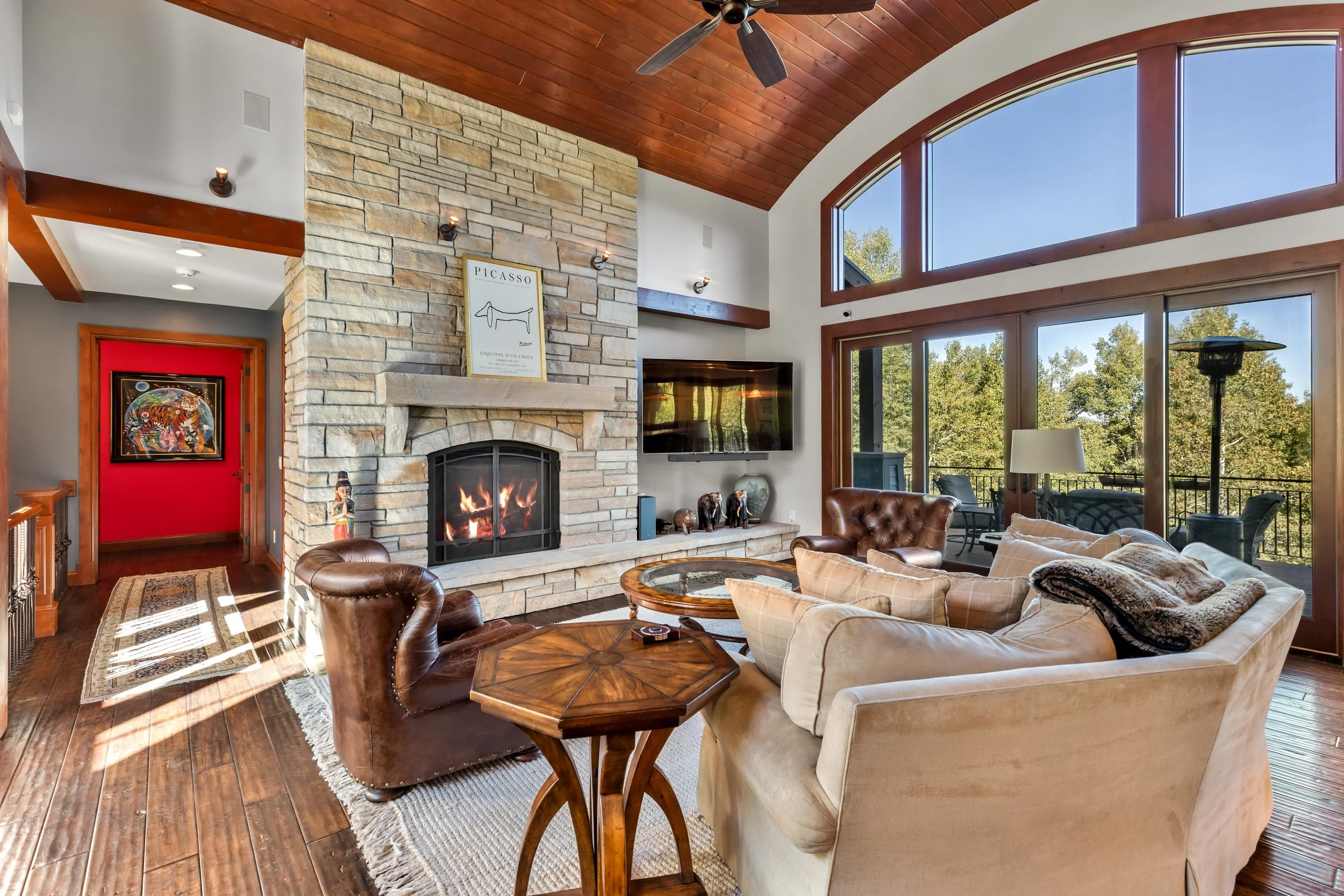 Living room with a stone fireplace, brown leather armchairs, a beige sofa with plush throws, a wooden coffee table, large windows showing trees outside, and a mounted flat-screen TV.