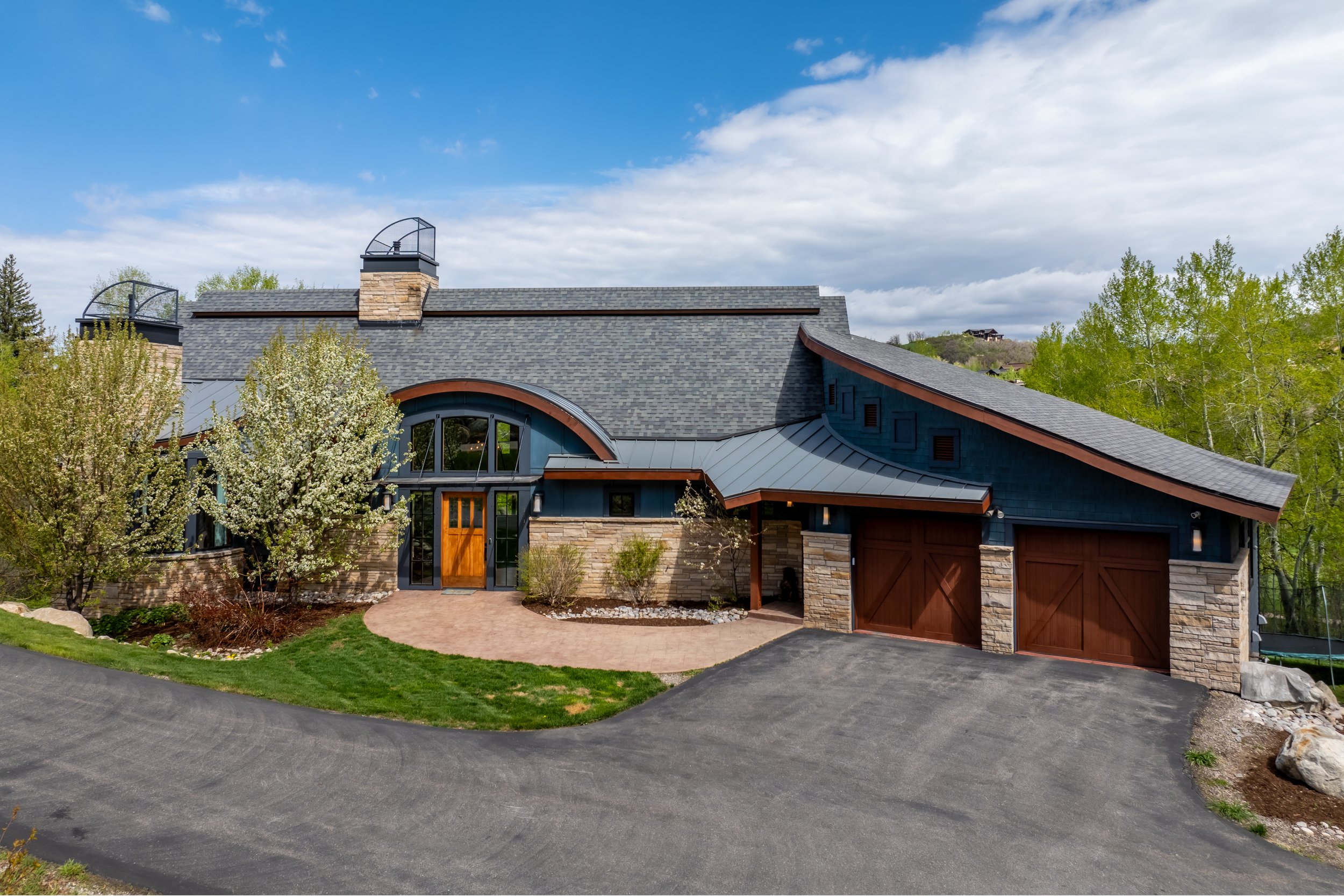 Modern house with stone accents and a metal roof, surrounded by green trees and blue sky.