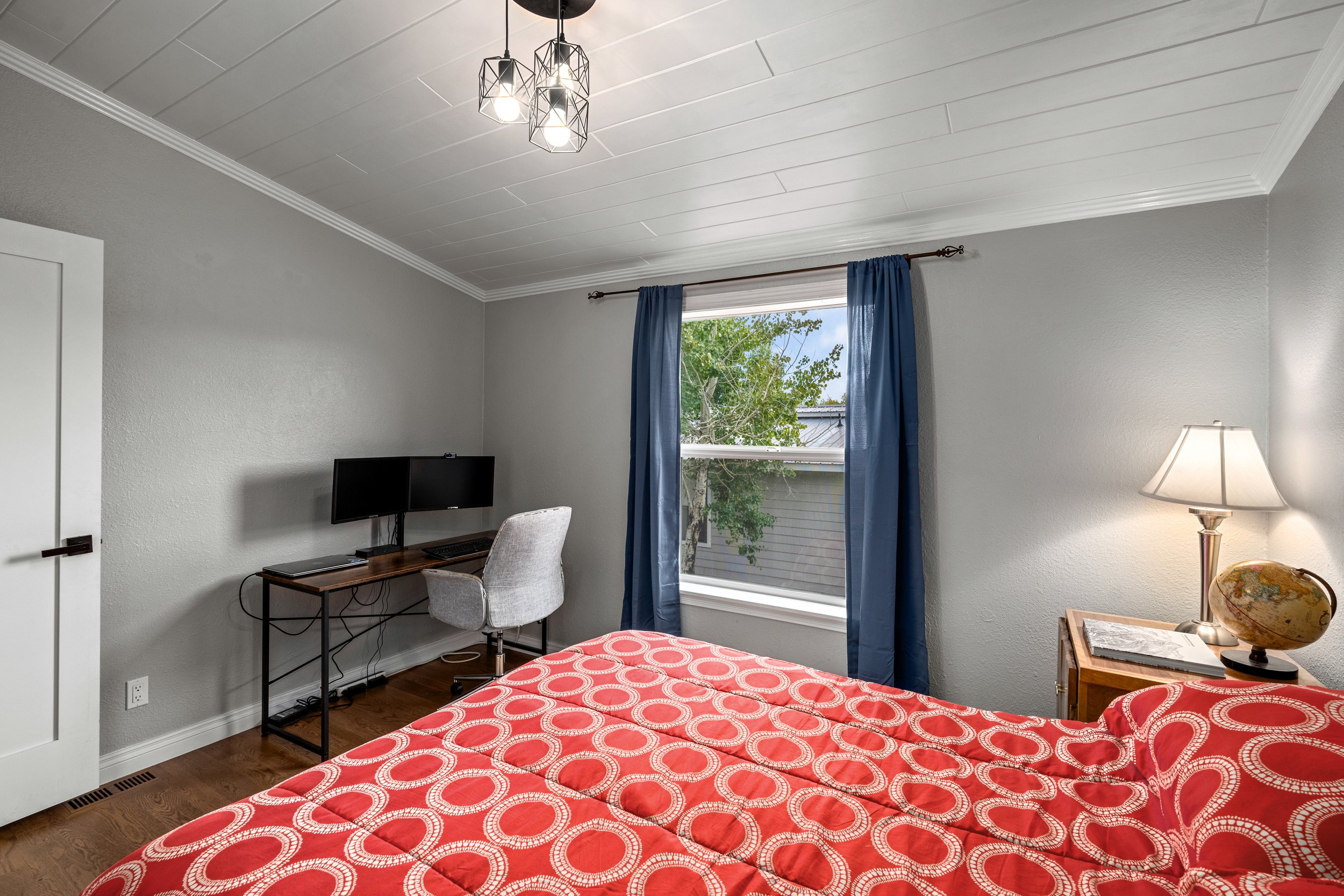 Small bedroom with gray walls, featuring a bed with a red and white circular patterned comforter, a wooden desk with a dual monitor setup, gray desk chair, and a bedside table with a lamp and globe. A window with blue curtains is in the background.