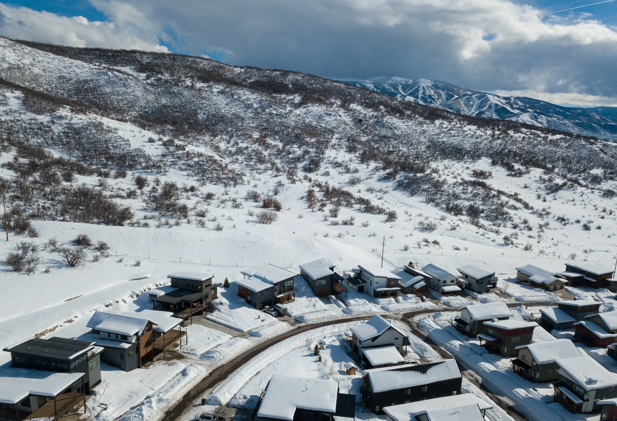 Aerial view of a snowy residential neighborhood at the base of wooded hills and mountains under cloudy skies.