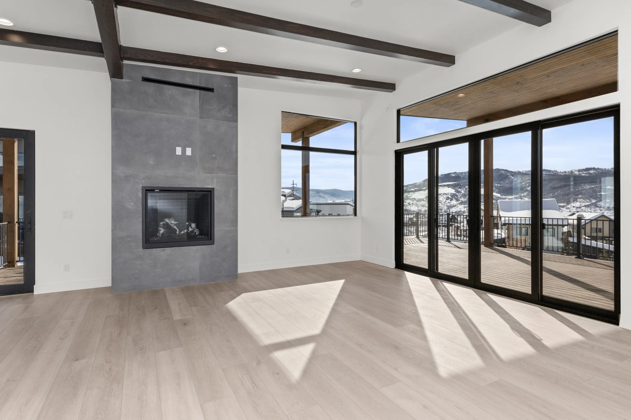 Modern living room with large windows, stone fireplace, wooden flooring, and outdoor deck view.