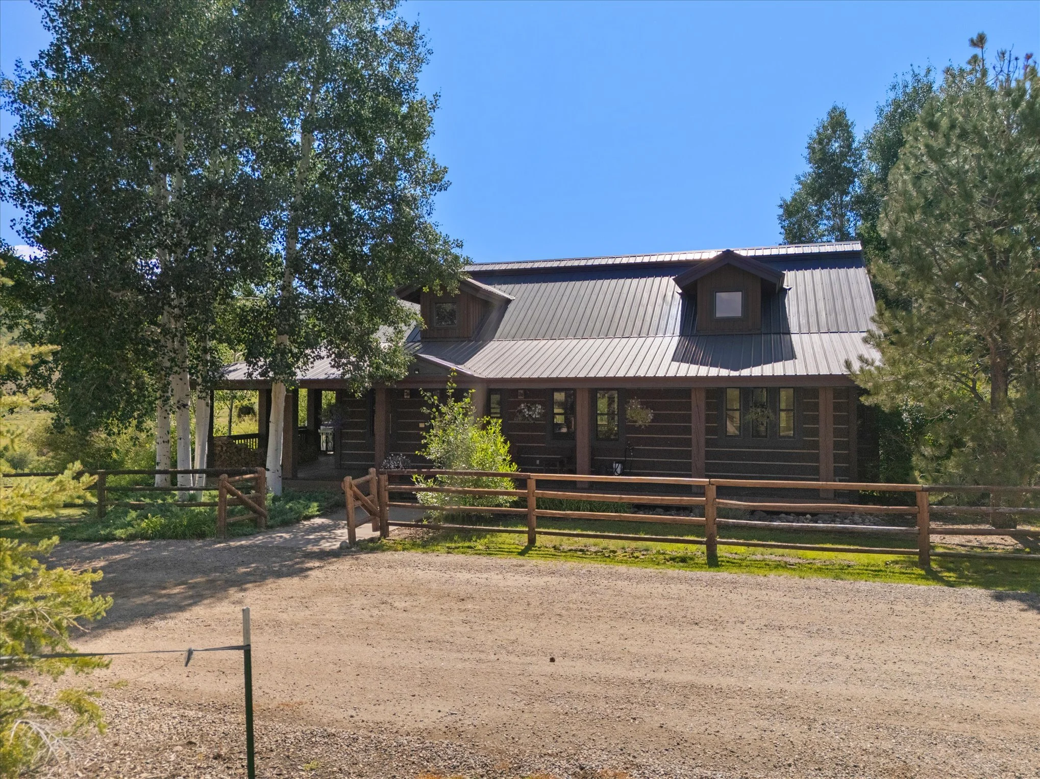 A rustic wooden house with a metal roof, surrounded by trees, with a gravel driveway and a wooden fence in front.