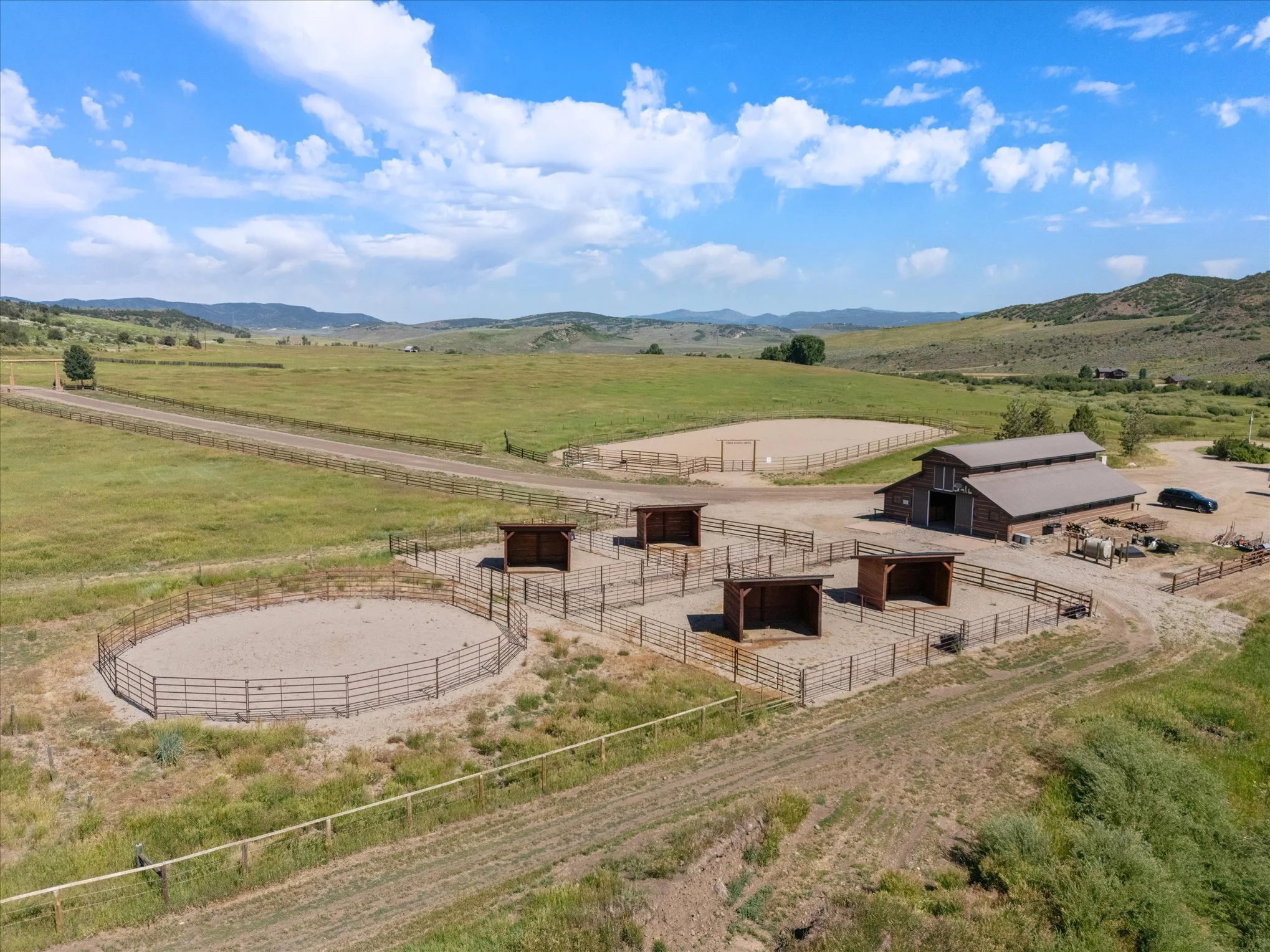 A horse riding arena with circular and rectangular enclosures, outdoor barns, and fencing, set in a rural landscape with rolling hills, green fields, and a partly cloudy sky.