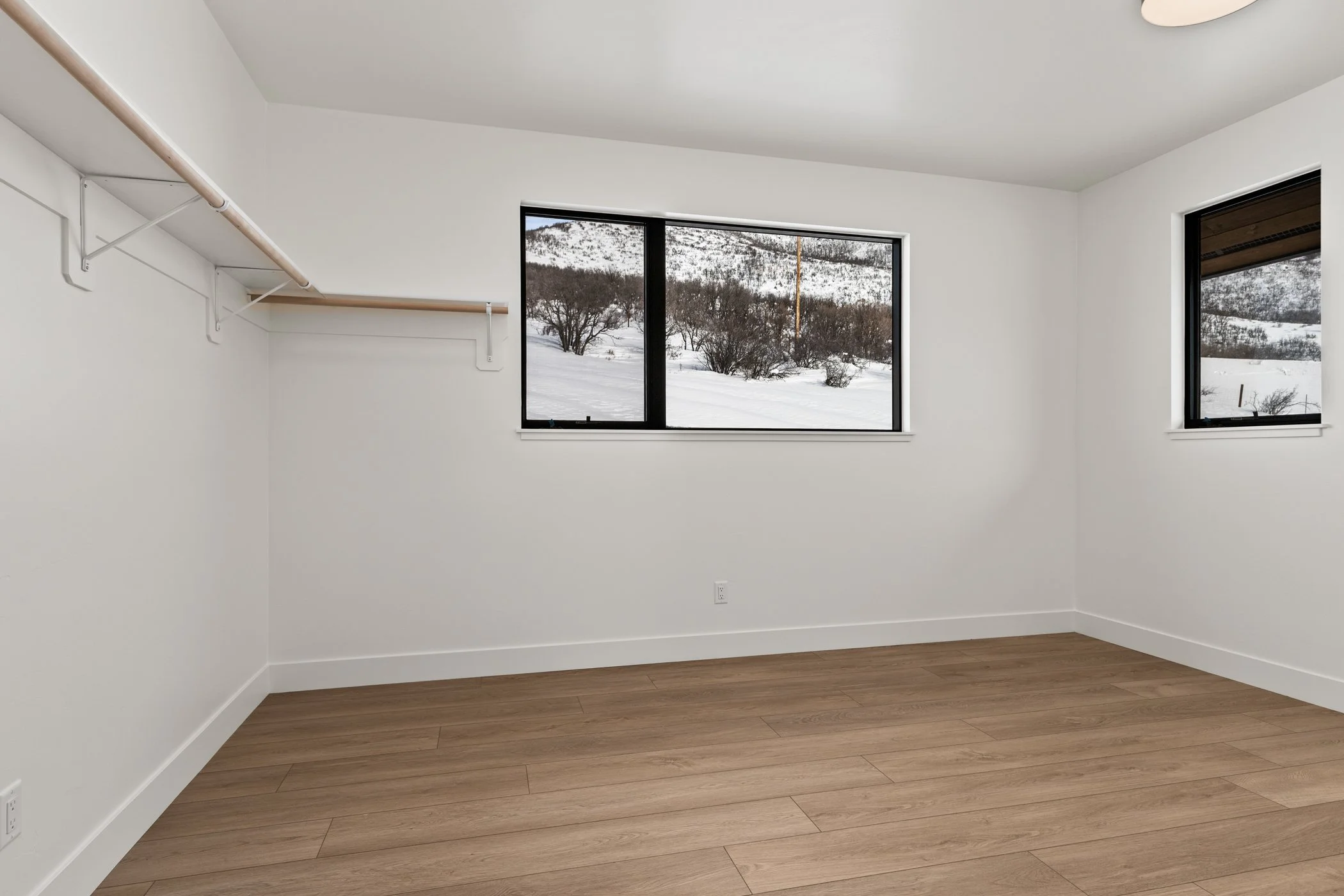 Empty room with light wood flooring, white walls, and two large windows displaying a snowy landscape outside. A closet rod without hangers is attached to the wall on the left.