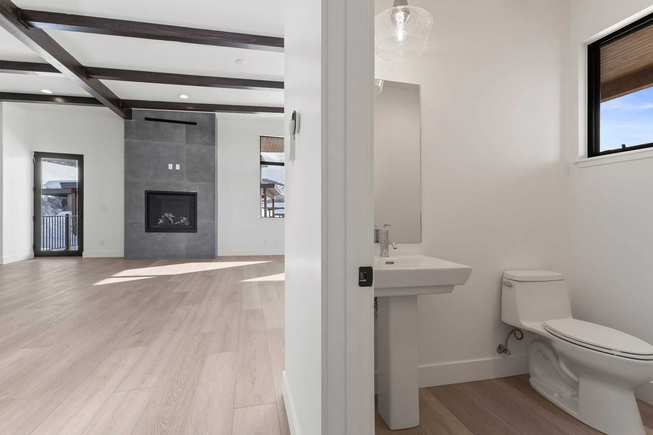Modern living room with wooden floors, a gray feature wall with a fireplace, exposed ceiling beams, and adjacent bathroom with a sink and toilet.