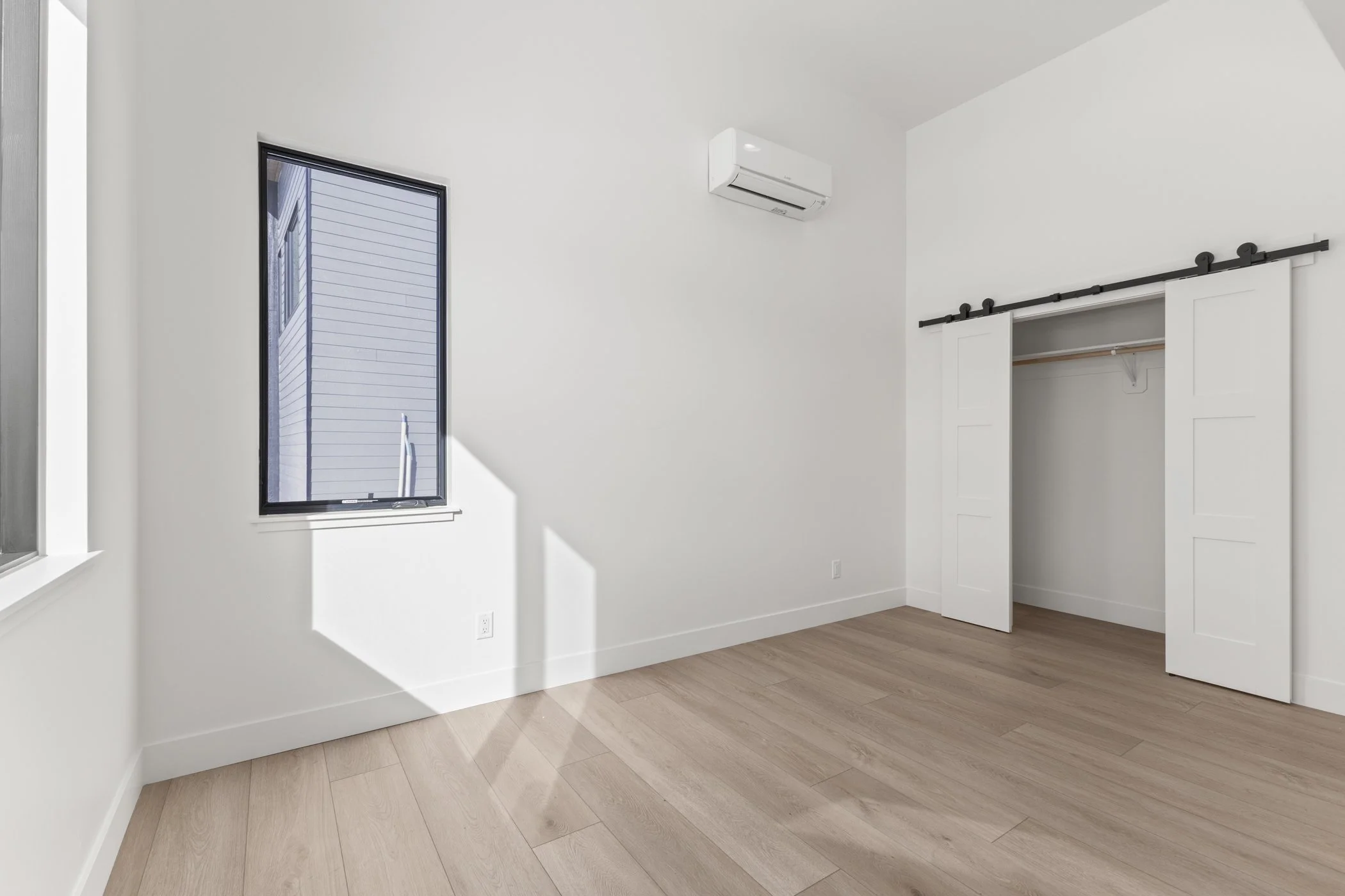 Minimalist empty room with wooden flooring, a window, an air conditioner, and a sliding closet door.