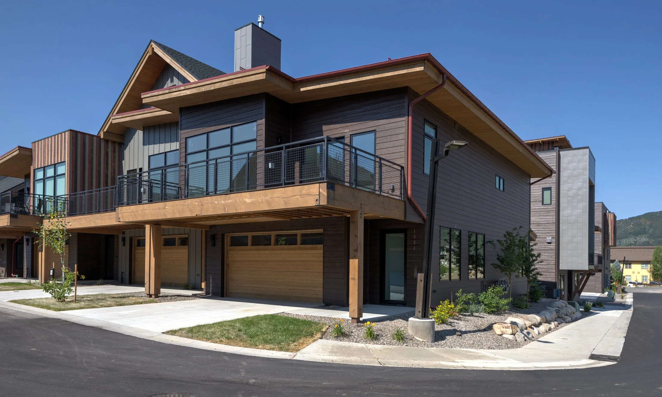 Modern townhouse with wooden accents and large windows, featuring an elevated deck and garage, situated on a paved street.