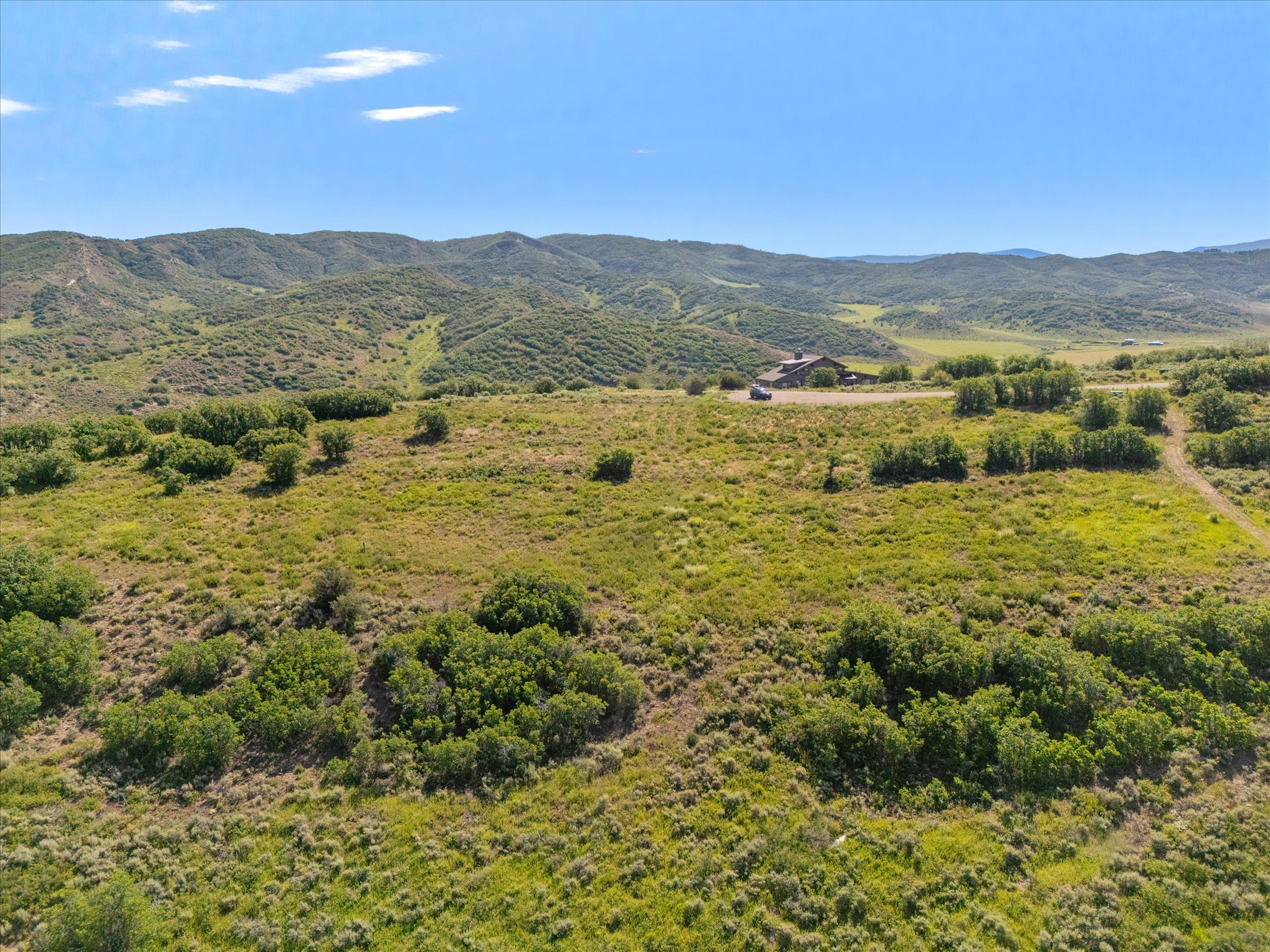 A scenic view of a hilly landscape with green vegetation, small trees, and a few buildings in the distance under a clear blue sky.
