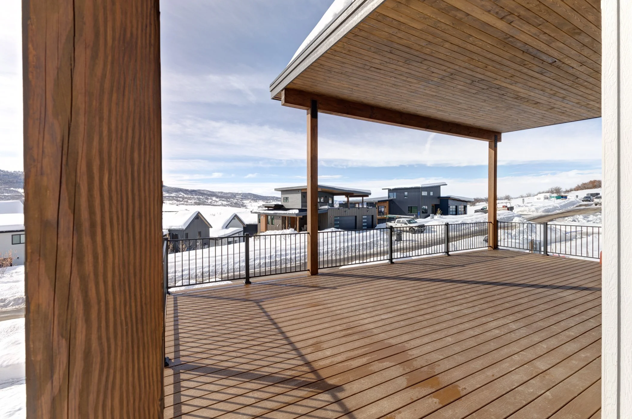 Covered wooden deck overlooking a snowy suburban neighborhood with modern houses and clear sky.