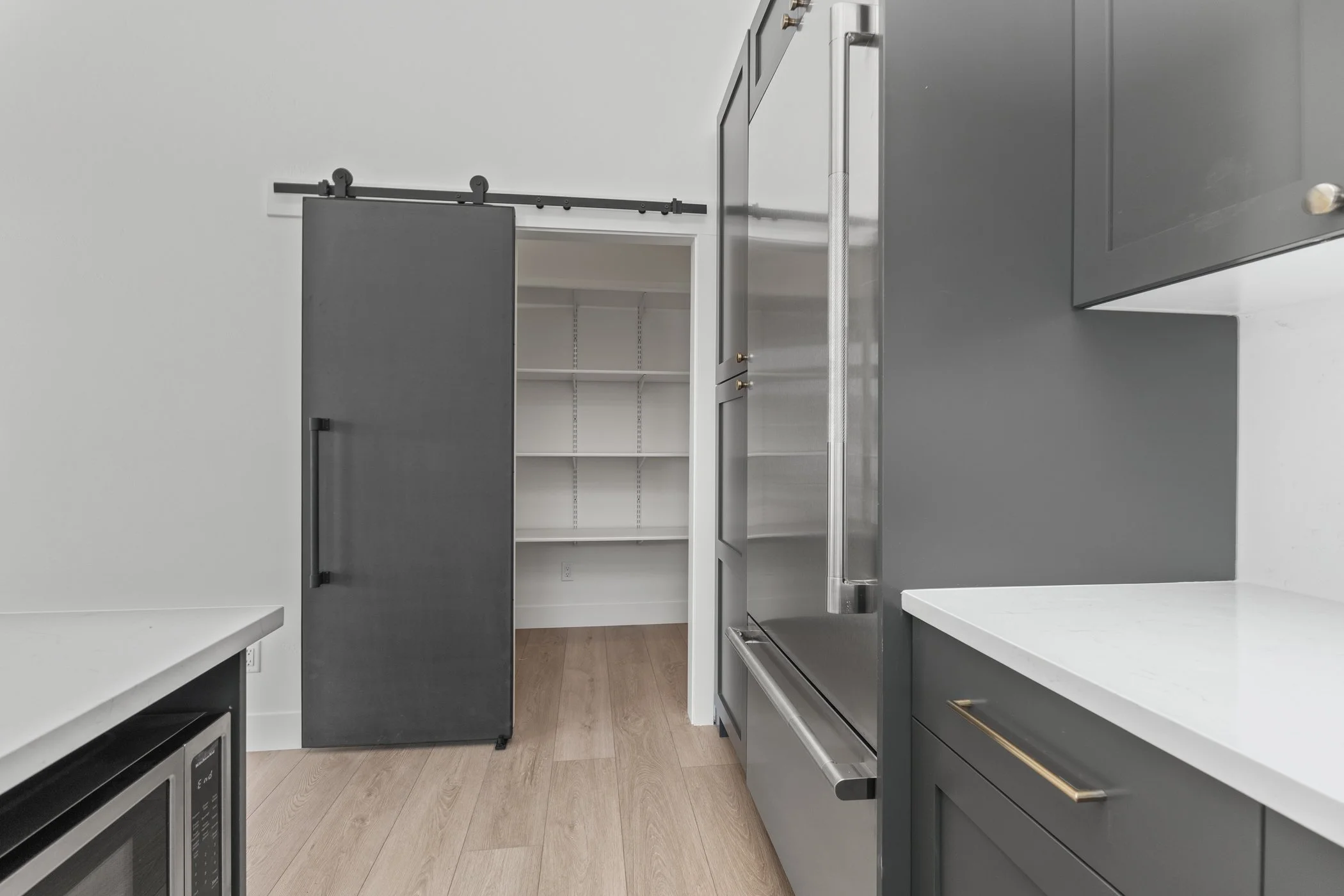 Modern kitchen with gray cabinets, sliding barn door, open pantry, stainless steel refrigerator, and light wooden flooring.