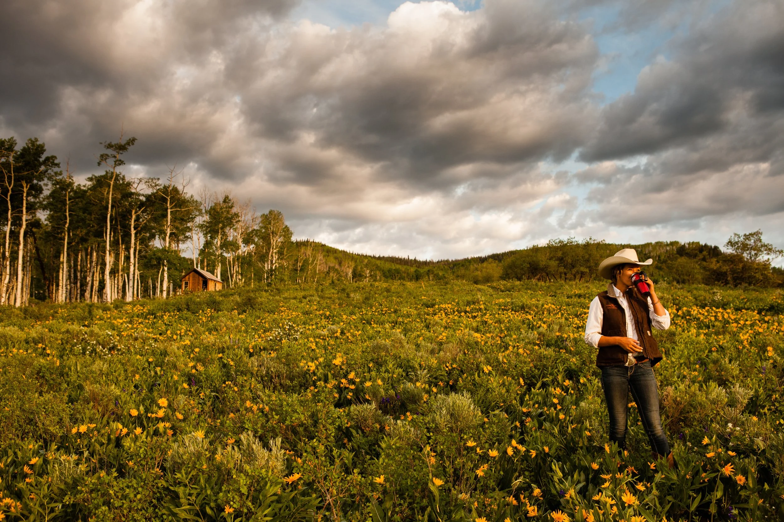 A woman wearing a cowboy hat, white shirt, brown vest, and jeans standing in a field of yellow flowers, drinking from a red cup, with a cloudy sky above and a small wooden cabin among trees in the background.
