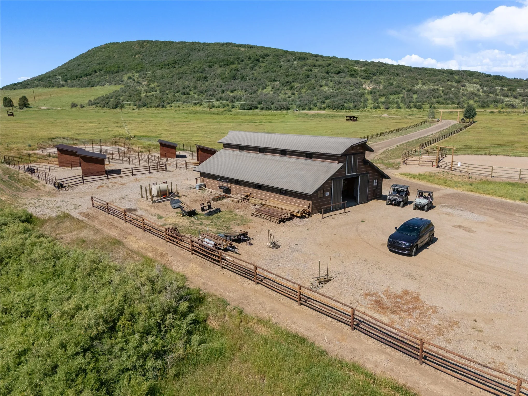 A large rural outdoor equestrian facility with a barn, horse stalls, fenced riding arenas, and open fields, surrounded by green hills under a blue sky.