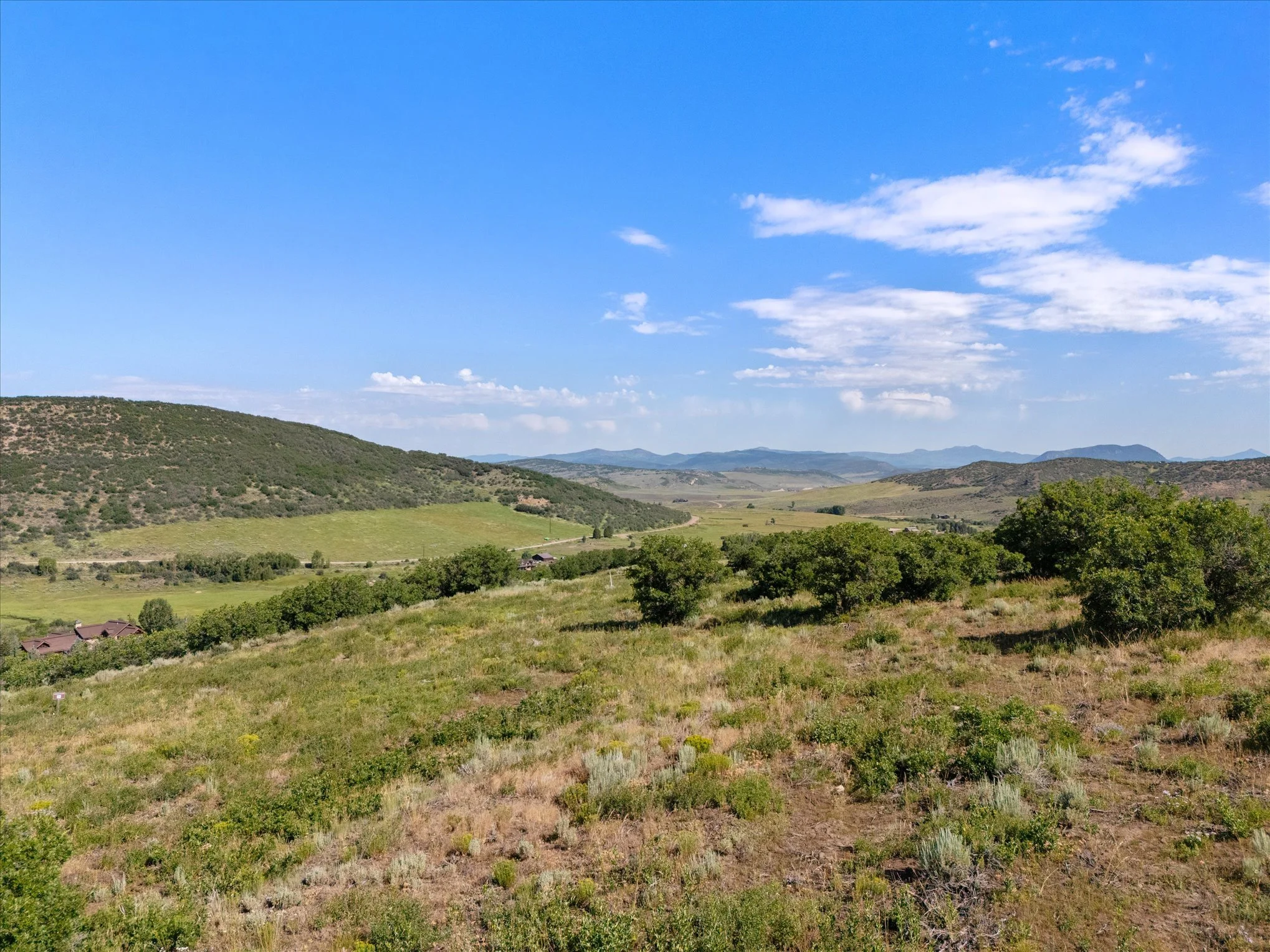 Scenic landscape of rolling green hills with scattered trees, some houses at the base, and a partly cloudy blue sky with distant mountain ranges.
