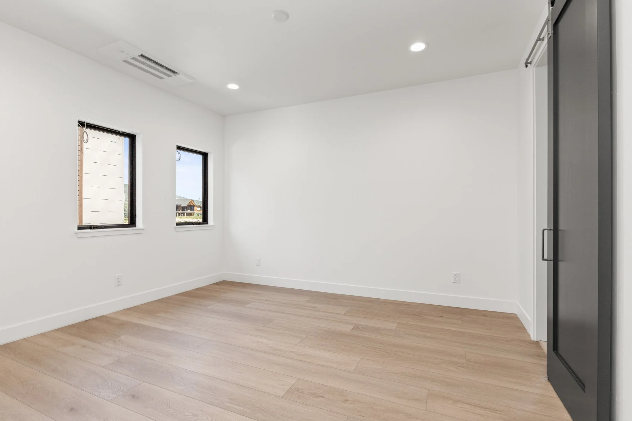 Empty white room with wooden floor and two windows, featuring a dark sliding door.