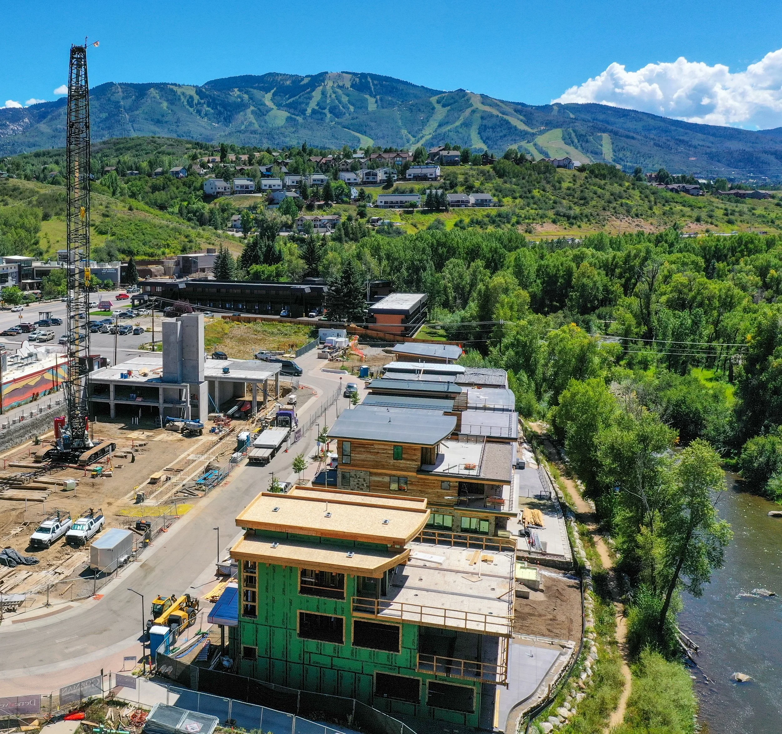 Aerial view of a construction site with several buildings in progress, featuring a large crane, vehicles, and adjacent greenery. In the background, there is a mountain with a ski resort and a residential area. A river runs adjacent to the constructio