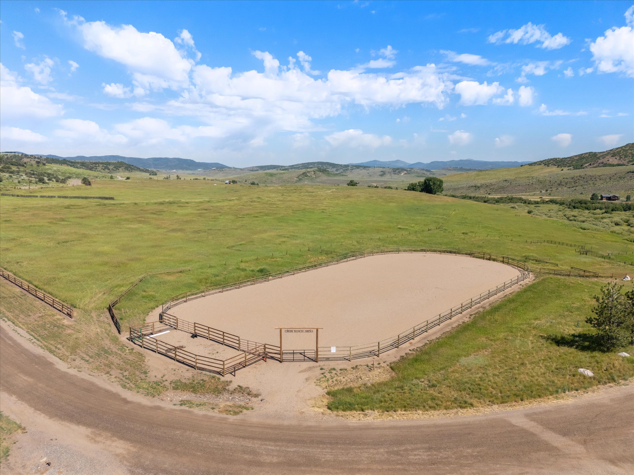 A fenced outdoor arena for horseback riding or training, located in a rural, grassy landscape with rolling hills and a blue sky with scattered clouds.