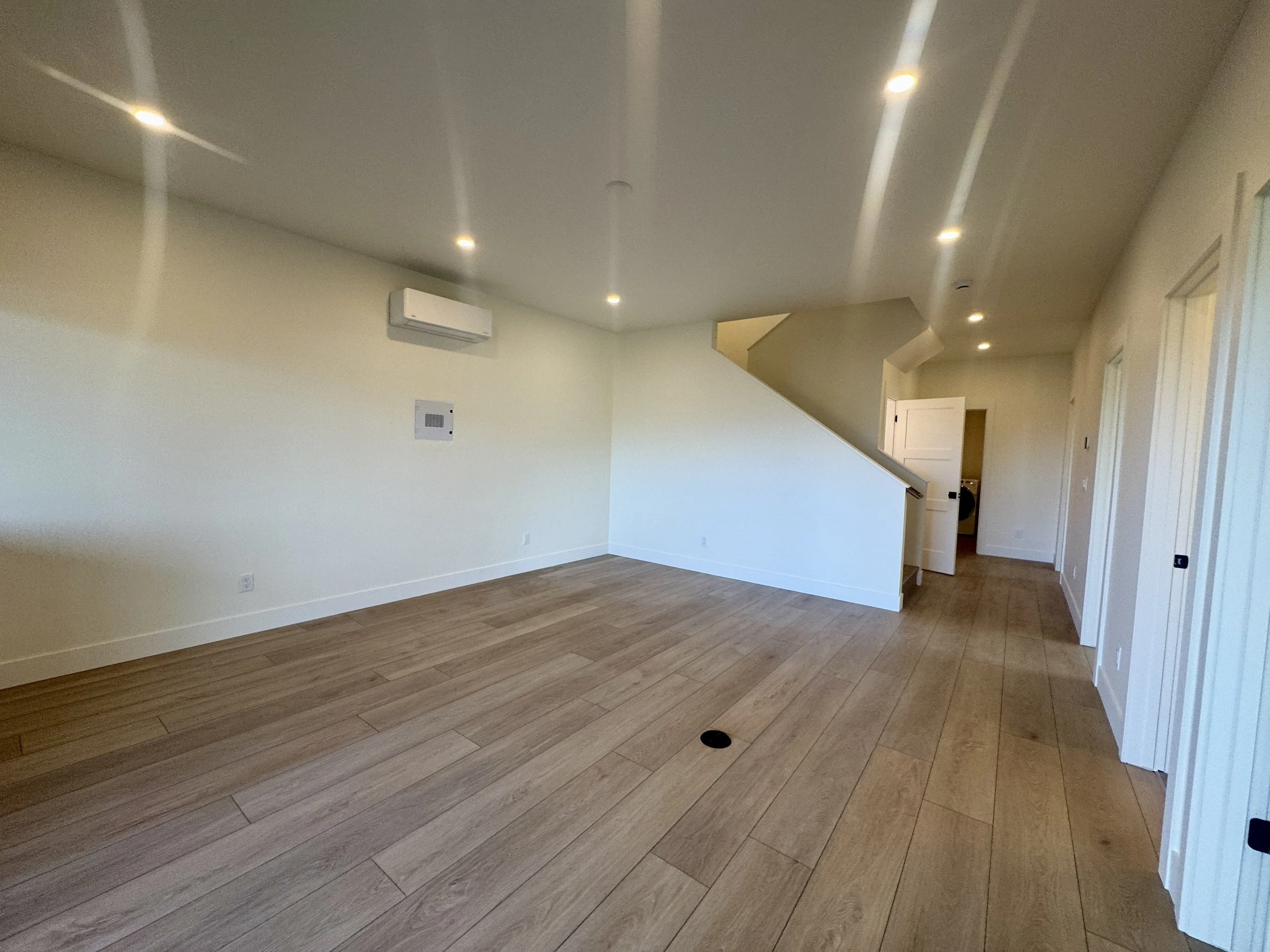 Empty living room with cream-colored walls, wood flooring, recessed ceiling lights, a wall-mounted air conditioner, and a staircase leading to the upper level.