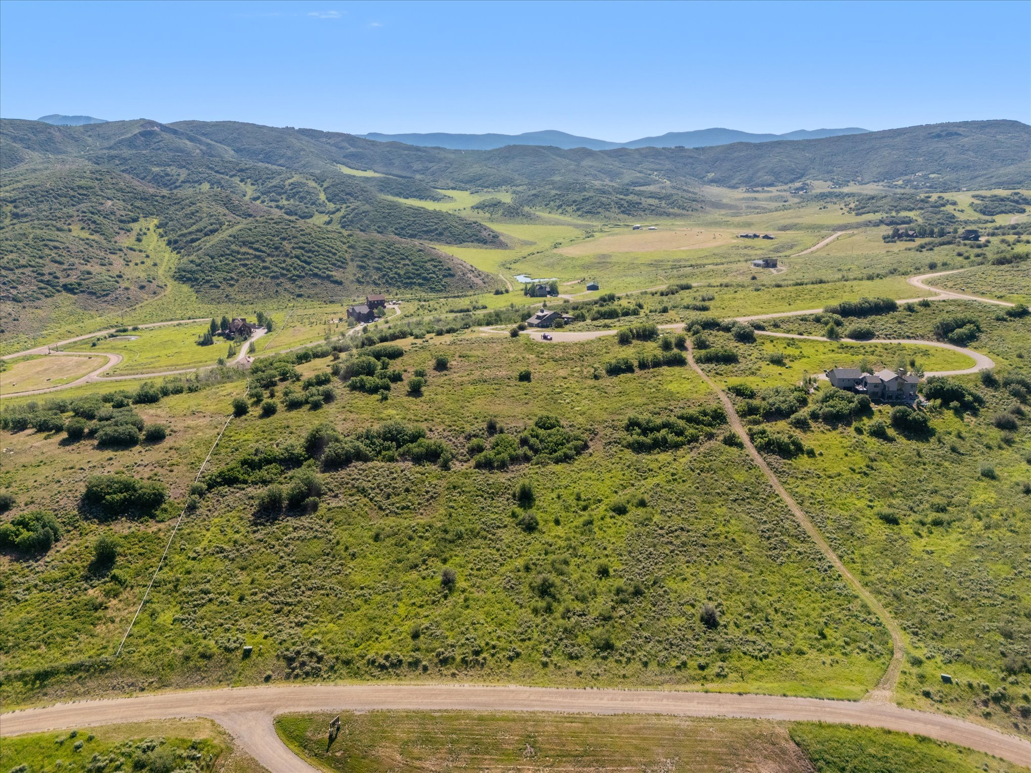Aerial view of a rural landscape with winding roads, scattered houses, green hills, and mountains in the background under a partly cloudy sky.