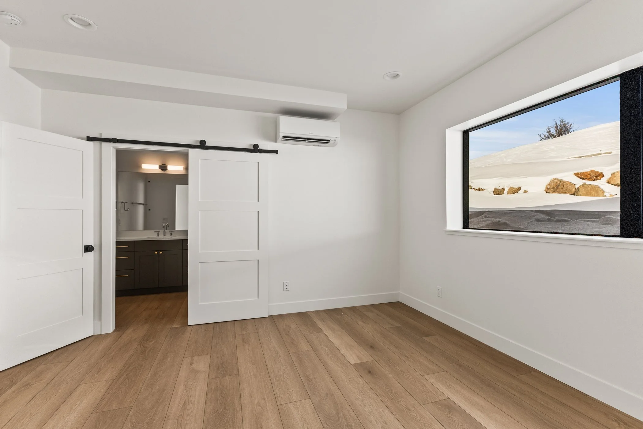 Minimalist room with wooden flooring, white walls, sliding barn door leading to a bathroom, large window overlooking snowy landscape, and wall-mounted air conditioning unit.