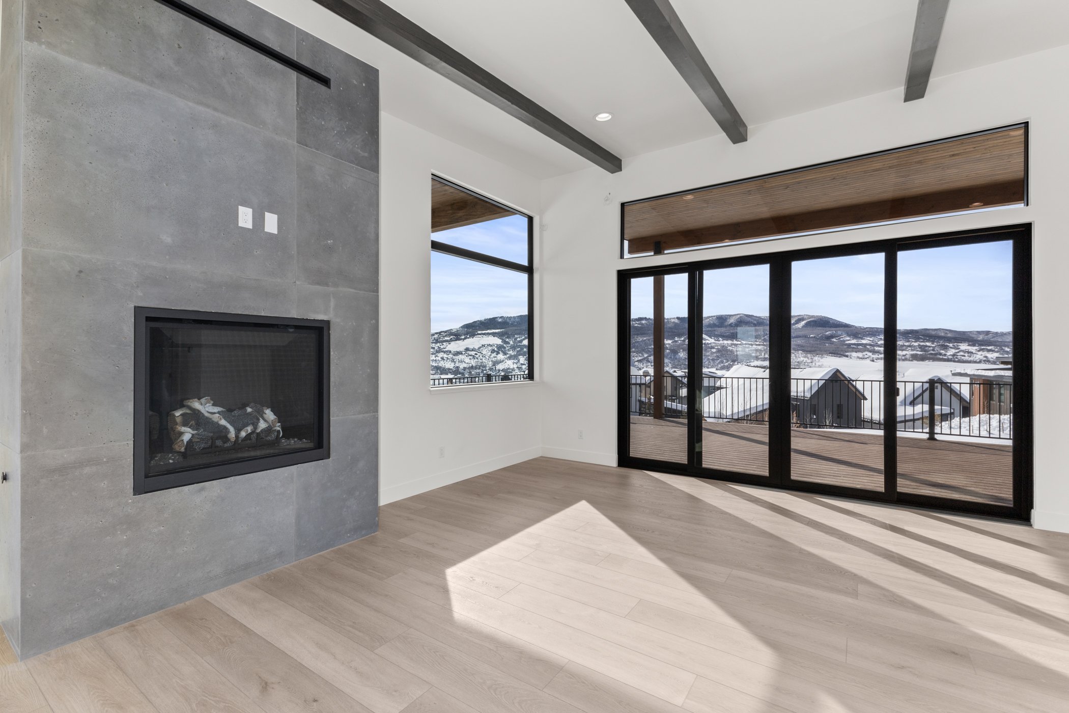 Modern living room with large windows, a concrete fireplace, wood floors, and a view of snow-covered mountains.