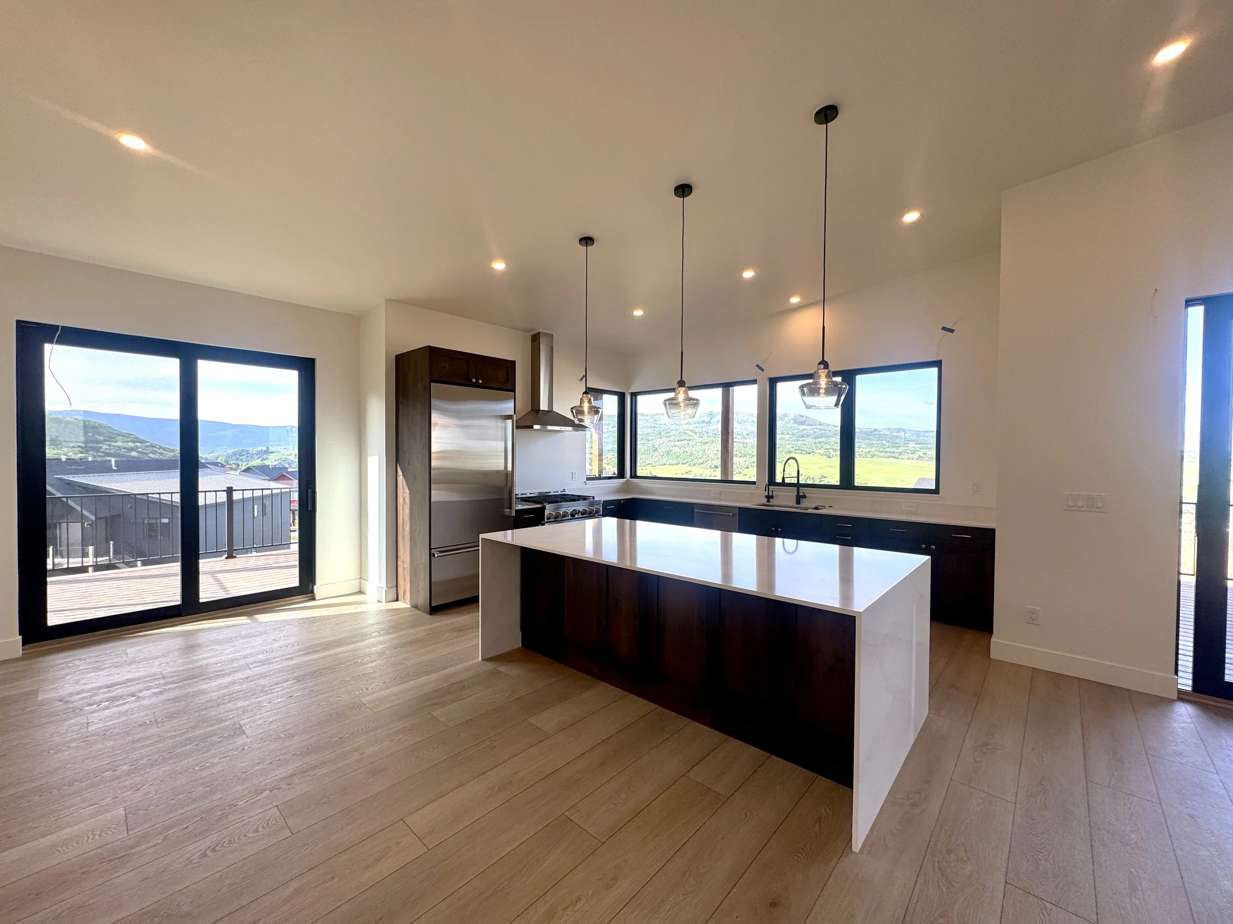 Modern kitchen with white island, dark cabinetry, stainless steel refrigerator, stove, and large windows showing scenic hills, with sliding glass door leading to a deck.