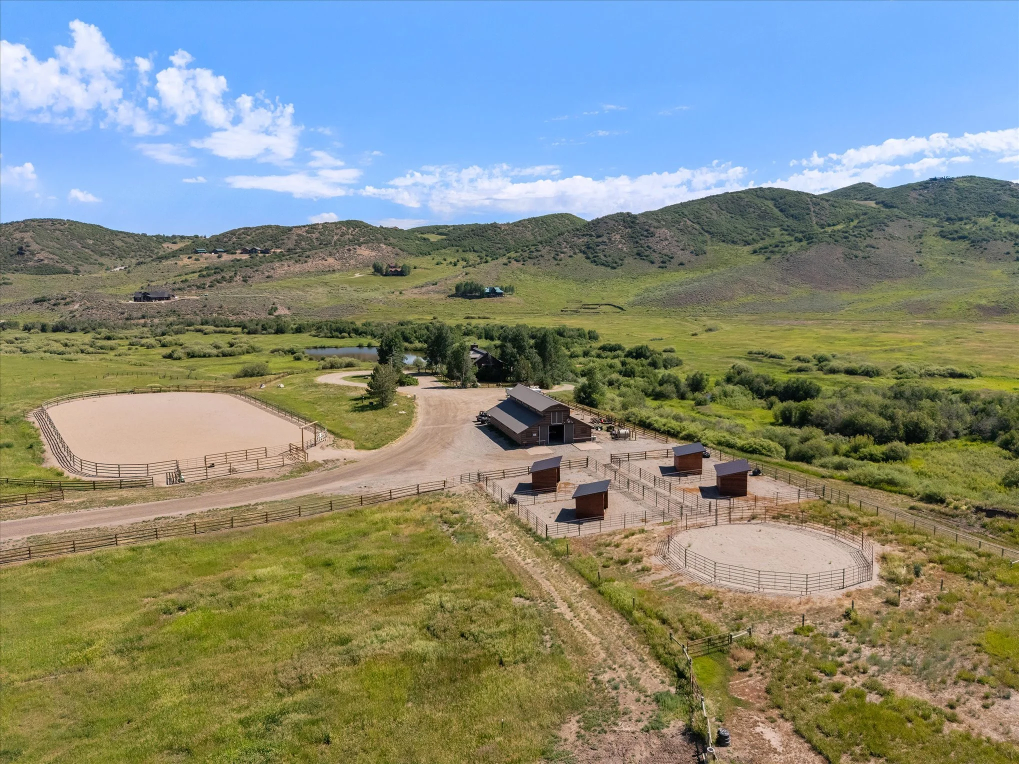 Aerial view of a rural equestrian facility with two horse riding arenas, stables, and rolling green hills in the background under a partly cloudy sky.