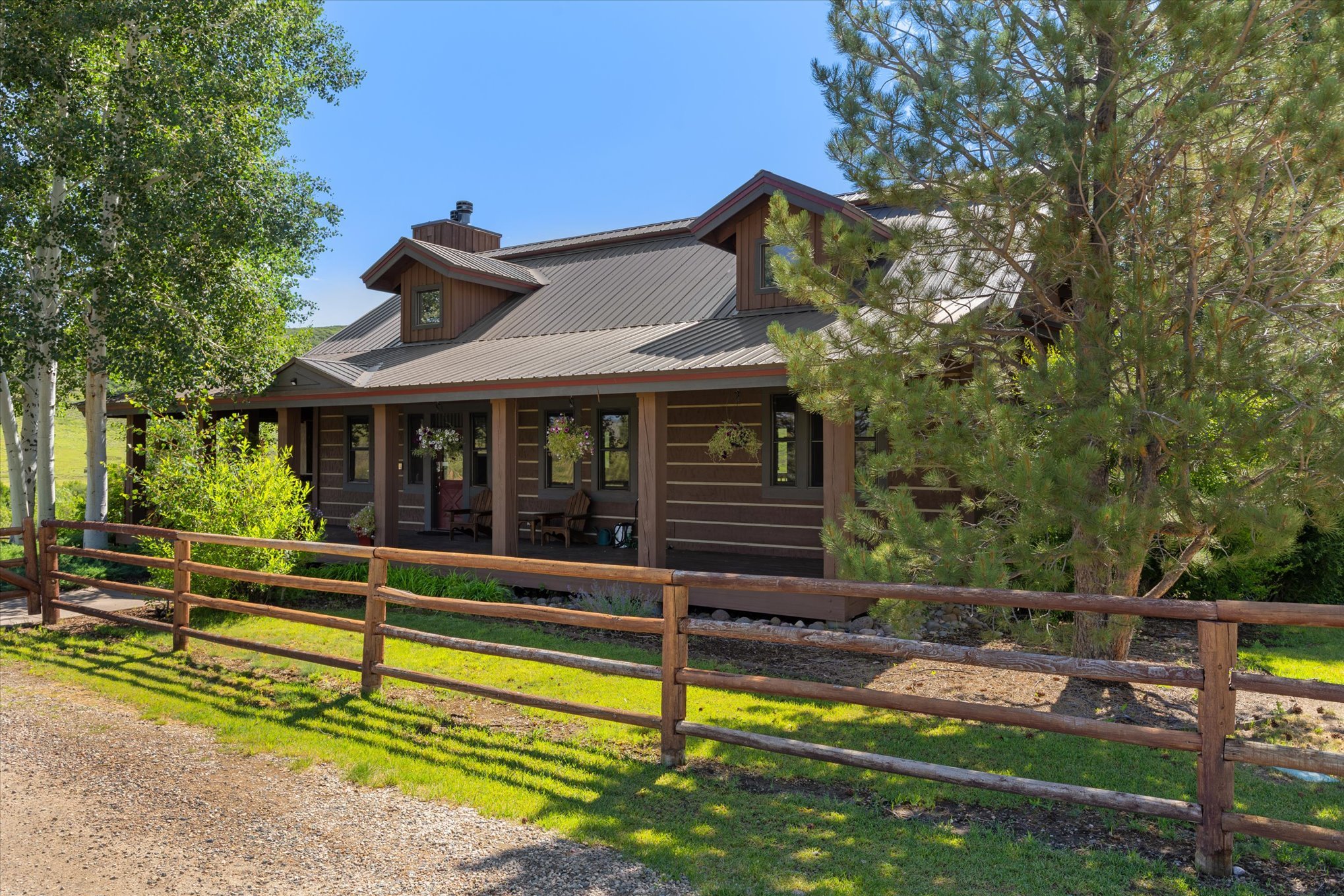 A wooden house with a metal roof, surrounded by trees, has a front porch with chairs and hanging flower baskets, enclosed by a wooden fence, on a sunny day.