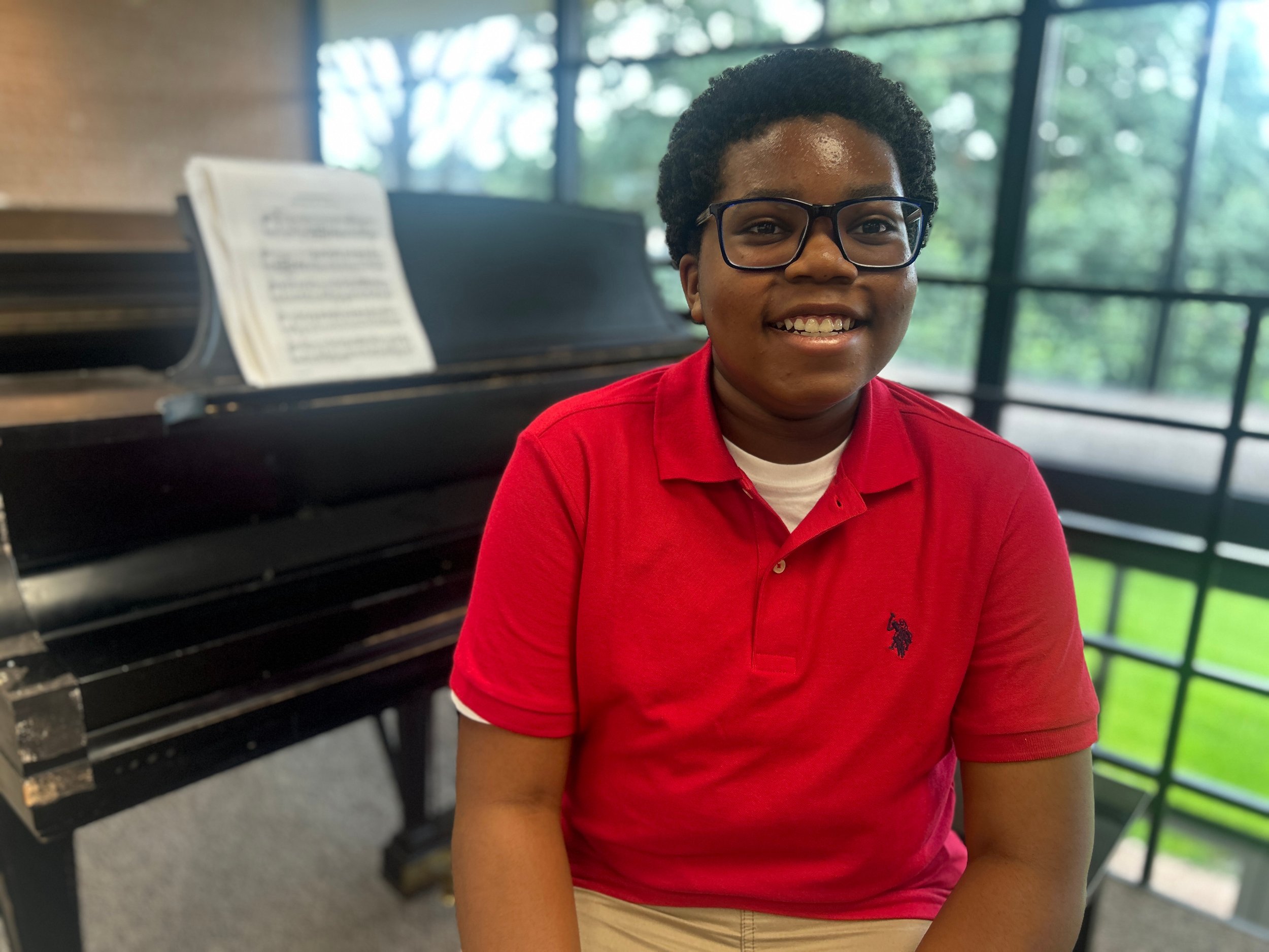A smiling young person with glasses wearing a red polo shirt, sitting in front of a black piano with sheet music, in a room with large windows overlooking green trees.