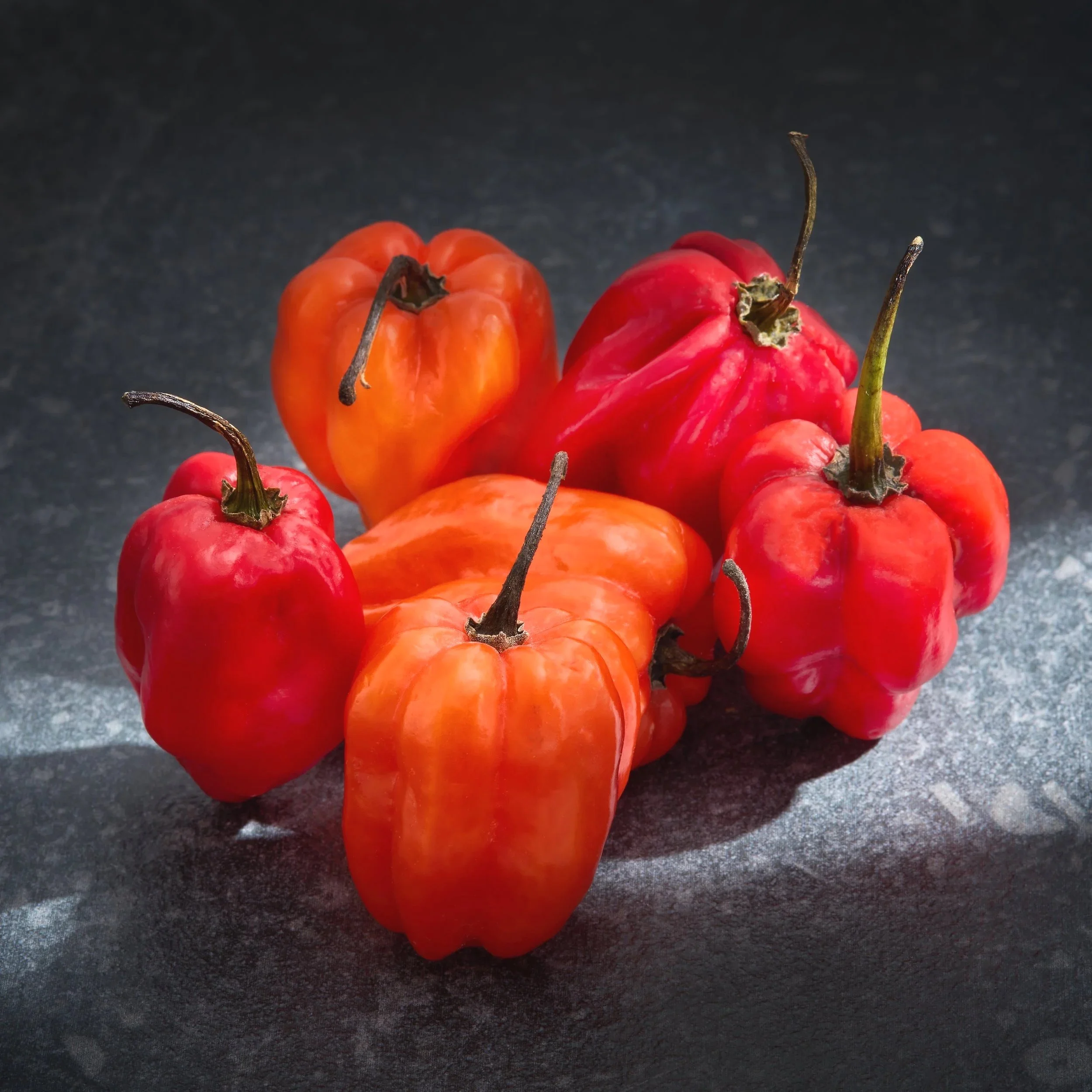 Close-up of red and orange habanero peppers with dry stems on a black surface.